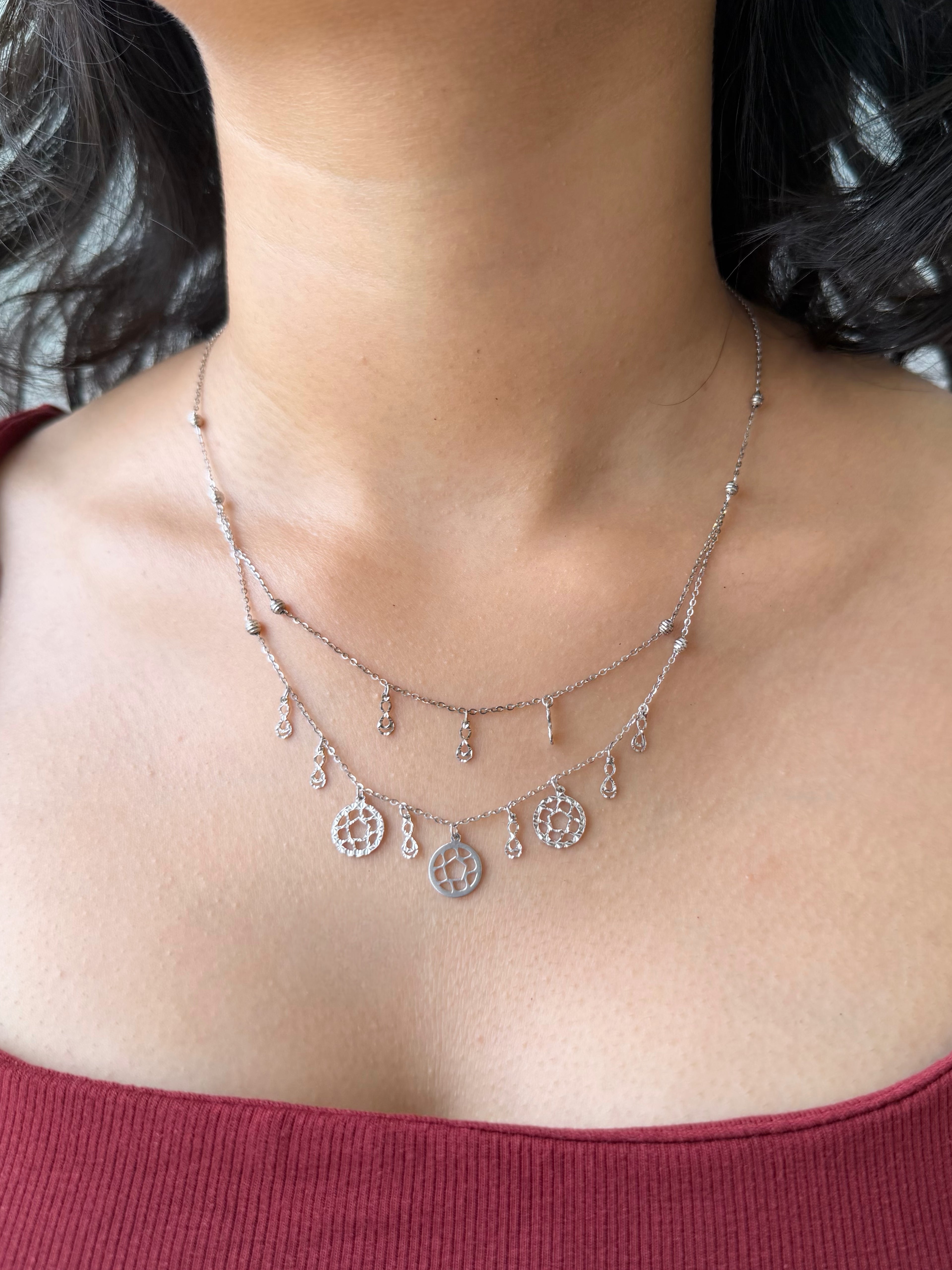 A close-up of a person's neck and upper chest wearing a layered silver necklace with small dangling charms and circular motifs.
