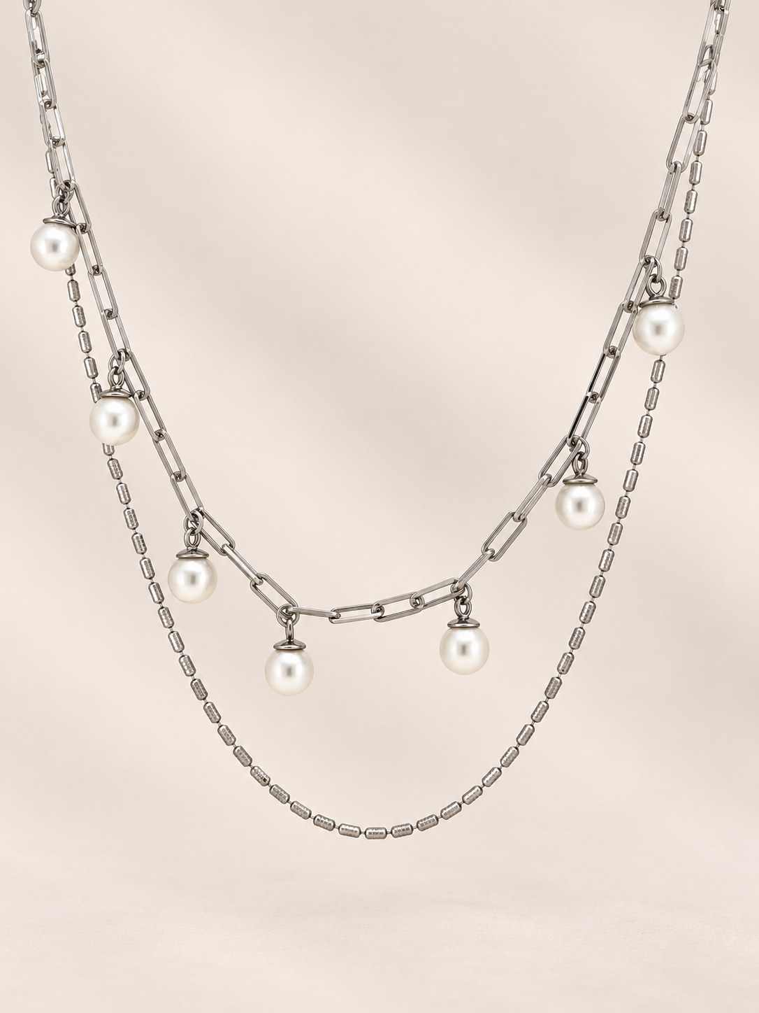 A layered silver-tone necklace featuring a paperclip chain, a fine chain, and five dangling white pearl beads, displayed against a soft beige background.
