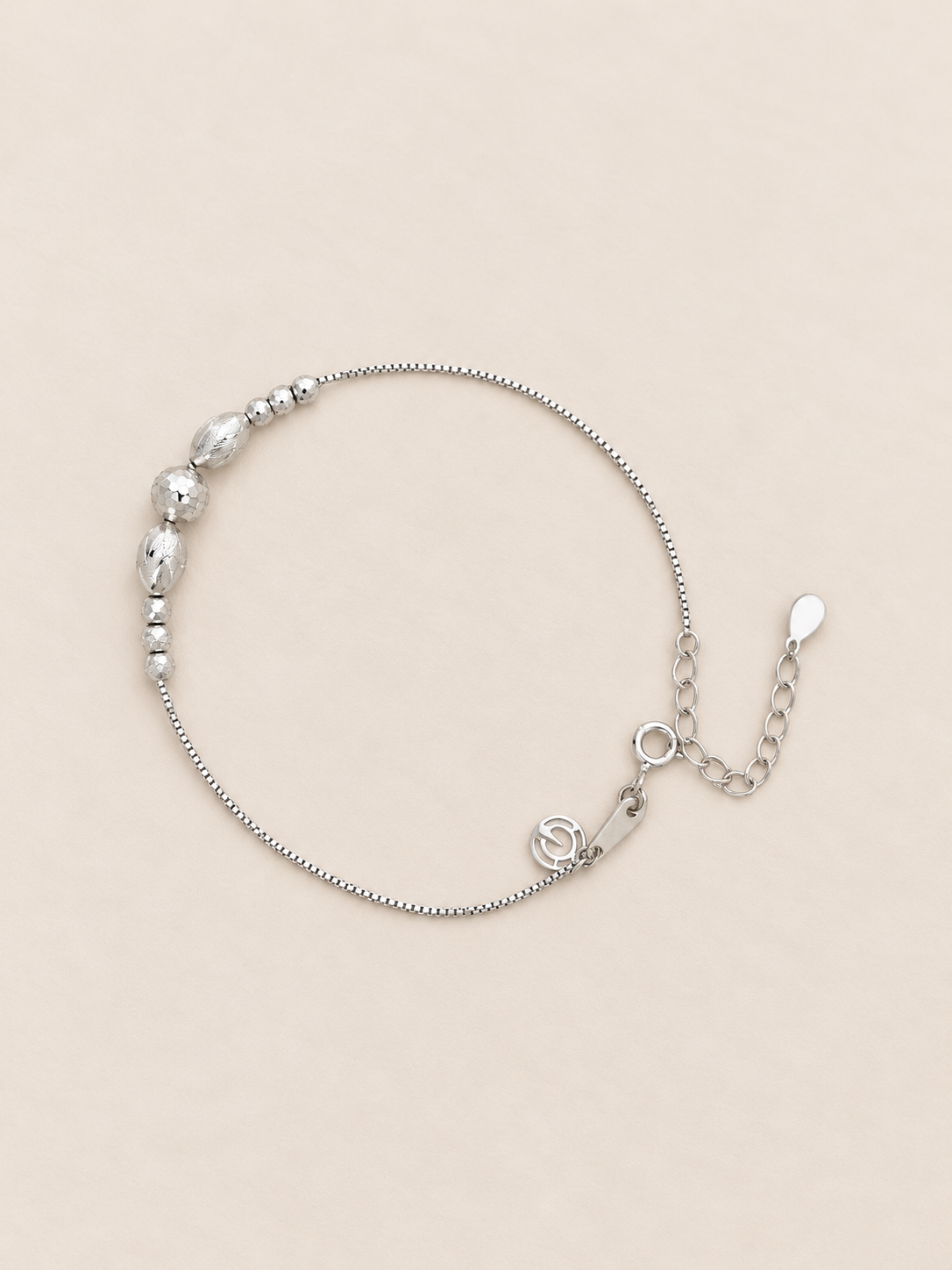 A delicate silver chain bracelet with a row of faceted silver beads on one side, a small silver charm, and an adjustable chain extender with a polished tip, displayed on a light beige background.