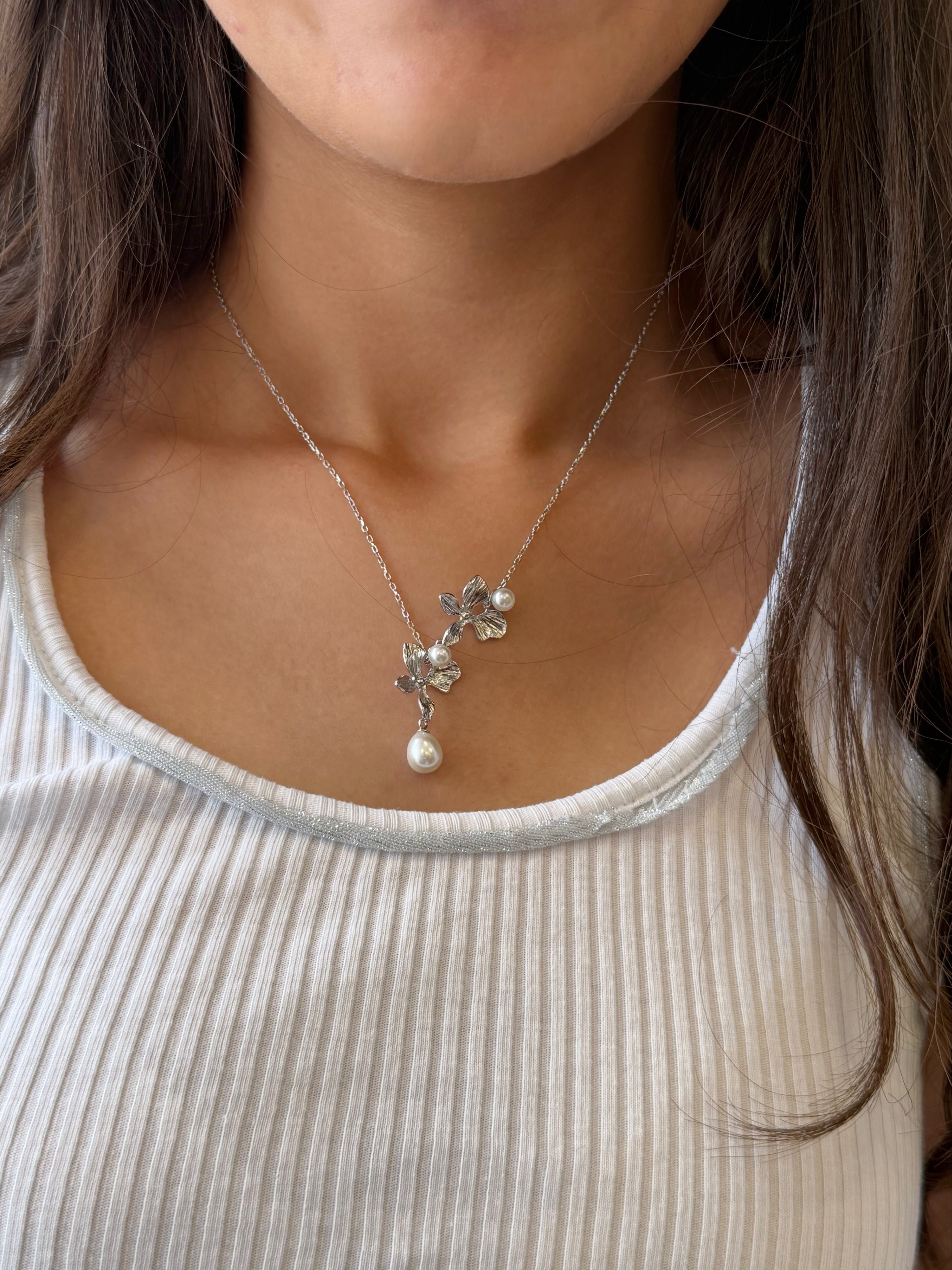 A close-up of a woman wearing a delicate silver necklace with a pearl pendant and small silver floral charms, styled with a ribbed white top.