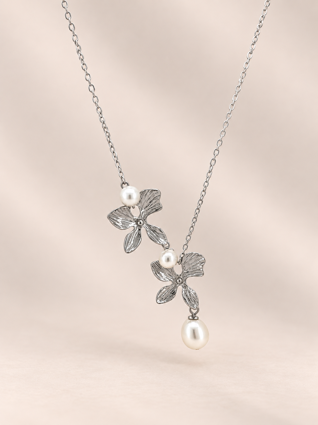 A silver-toned floral necklace with three textured flower motifs, accented by small white pearls and a larger dangling pearl, shown on a soft beige background.