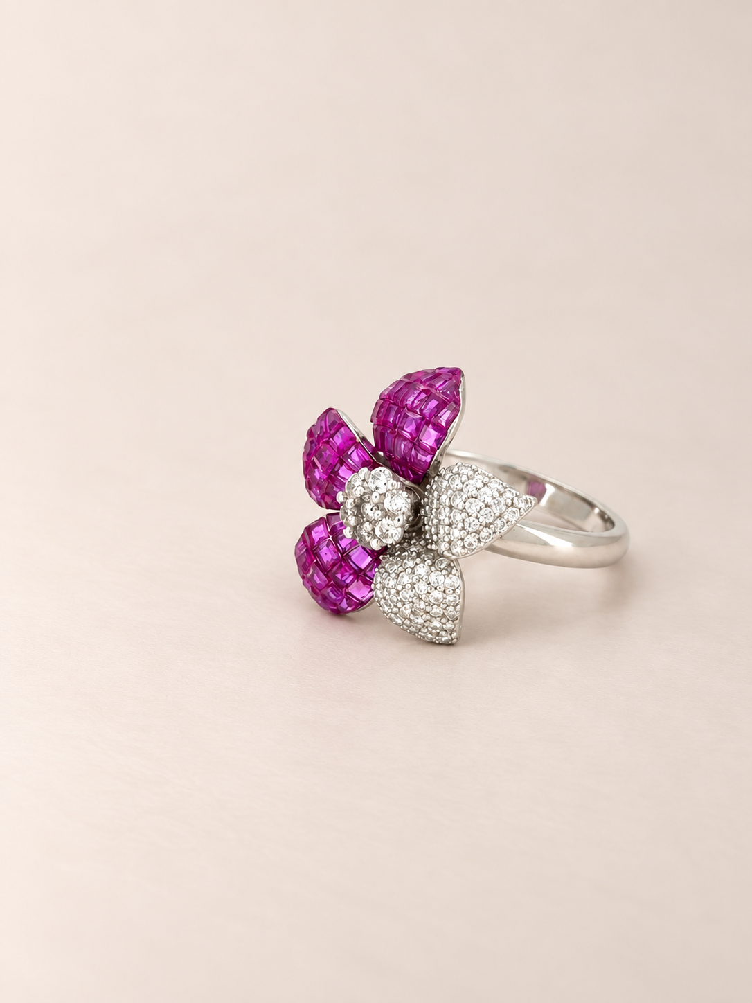 A silver flower ring featuring two bright magenta-pink textured petals and two pavé-set clear stones, photographed on a soft beige background.