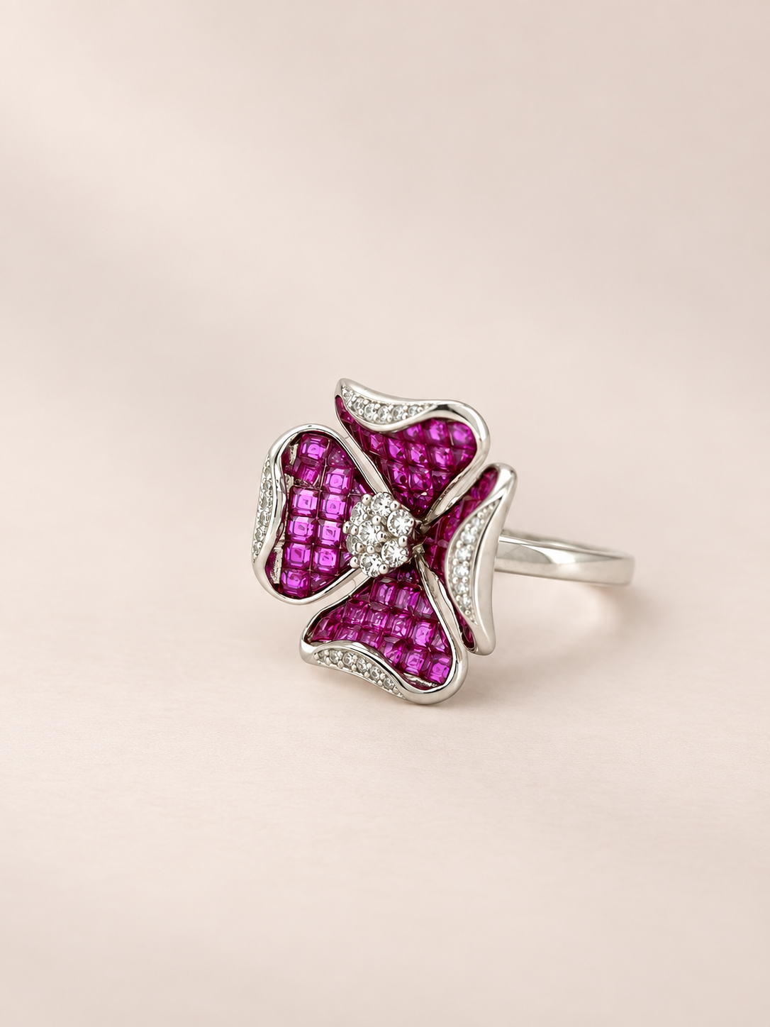 A silver ring featuring a four-petal flower design paved with bright pink gemstones, accented by a central clear stone and small pavé stones along the edges, set against a soft beige background.