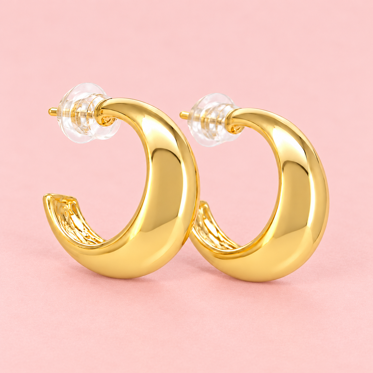 Polished gold-tone chunky crescent hoop earrings with a smooth, glossy finish and minimalist curved design displayed against a pink background.