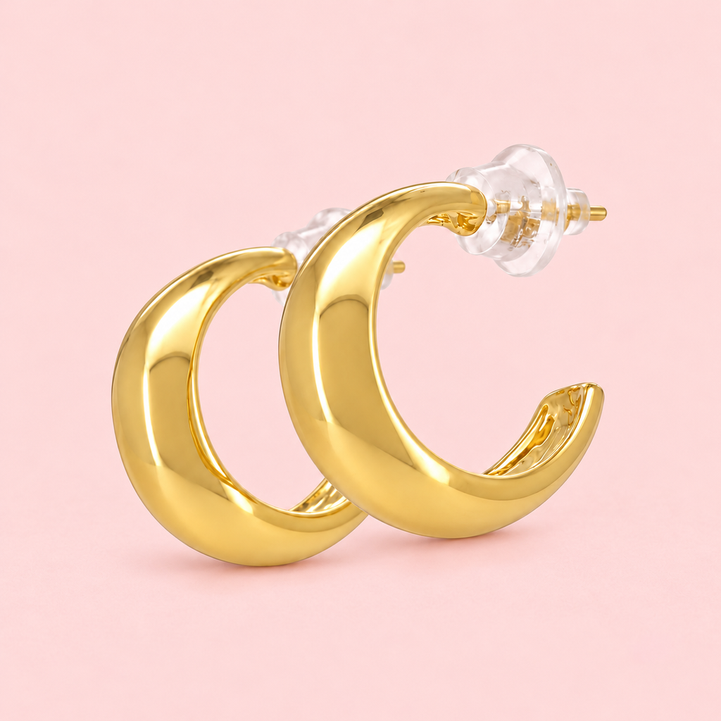 Chunky gold-tone hoop earrings with a rounded crescent shape and polished finish displayed on a pink background.