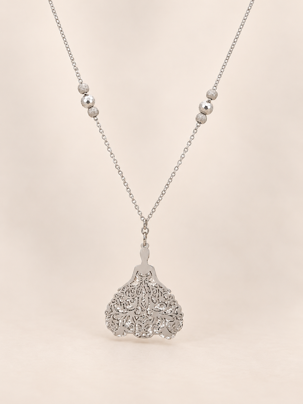 A silver pendant necklace featuring a detailed princess pendant and small silver beads on the chain, set against a solid pale pink background.