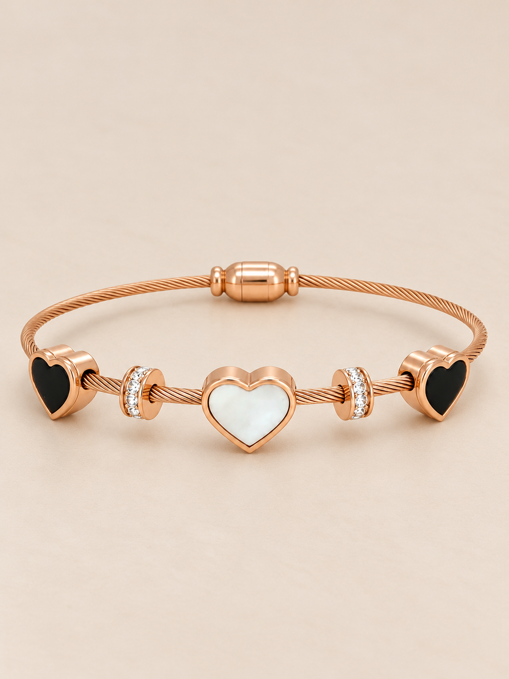 Rose gold stainless steel bracelet featuring three heart charms with black and white inlays and sparkling stone accents on a flexible wire band against a pink background. 