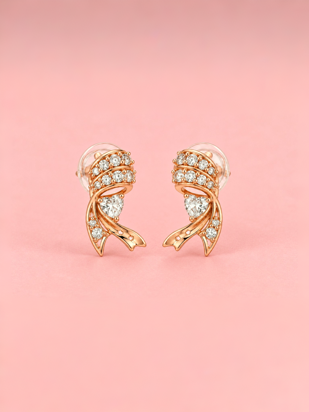 Rose gold-tone ribbon heart stud earrings featuring sparkling clear stones, heart-shaped crystal accents and intricate curved metal detailing displayed against a pink  background.