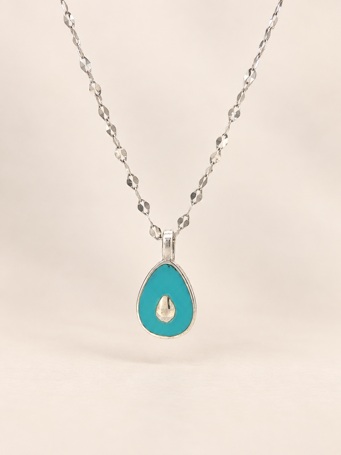 A silver teardrop pendant necklace featuring a vibrant turquoise blue enamel body with a small gold teardrop in the center, suspended from a silver faceted chain against a soft beige background.