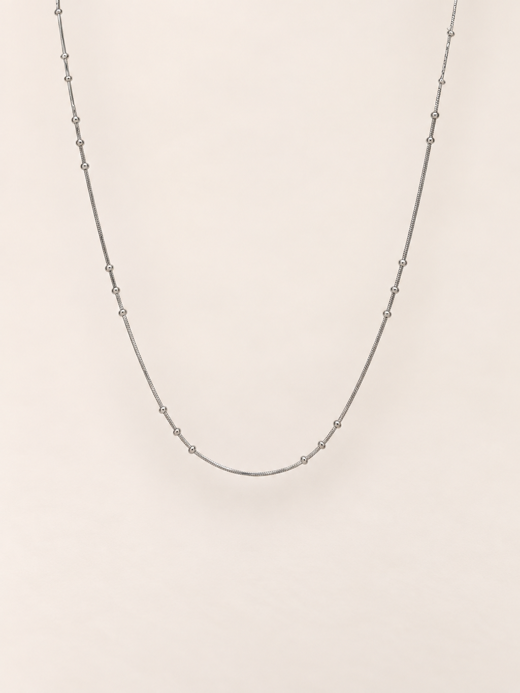 A delicate silver chain necklace with small, evenly spaced metallic beads, displayed against a solid, warm off-white background.