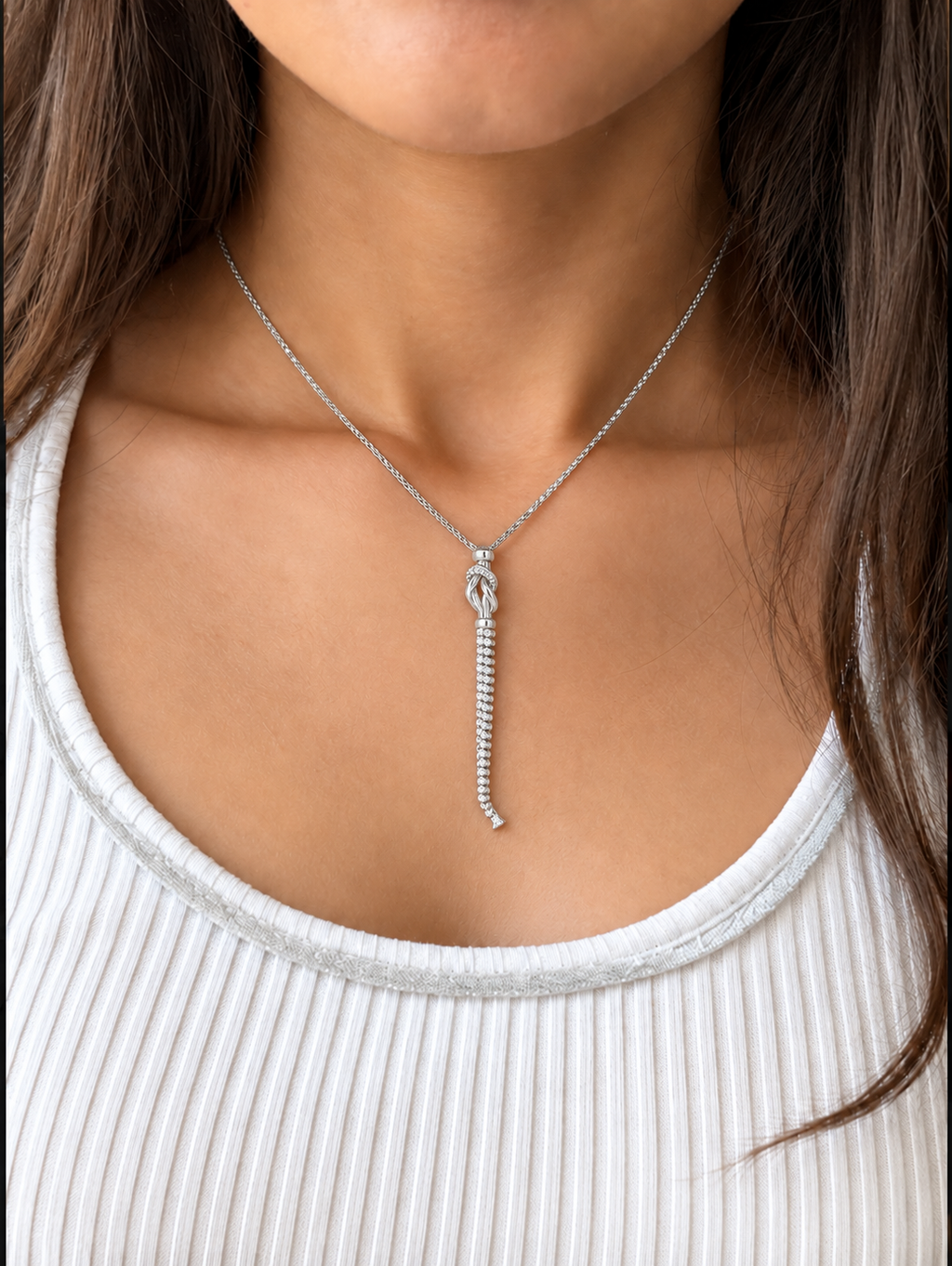 A silver-toned necklace featuring a delicate chain and a pendant with a knot-like top and a long, snake-like chain dangle, worn by a model on a white top.
