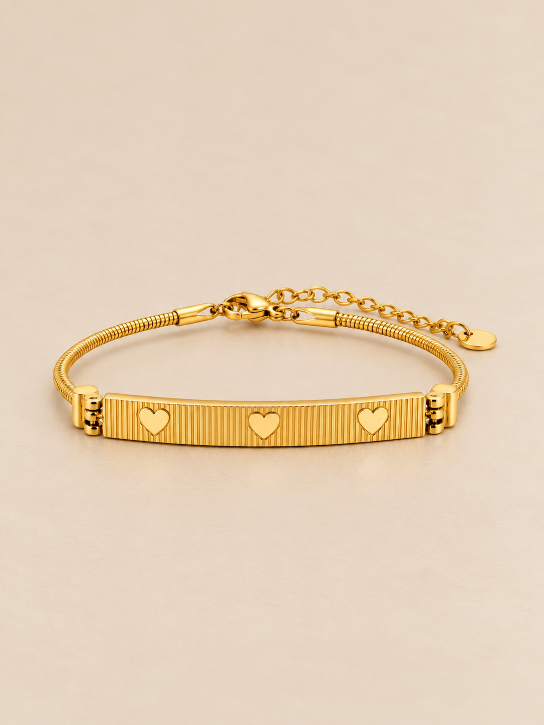 Gold-toned stainless steel bracelet featuring a textured rectangular bar with three engraved heart motifs.
Slim snake-chain bracelet with an adjustable extension, displayed on a clean pink background.