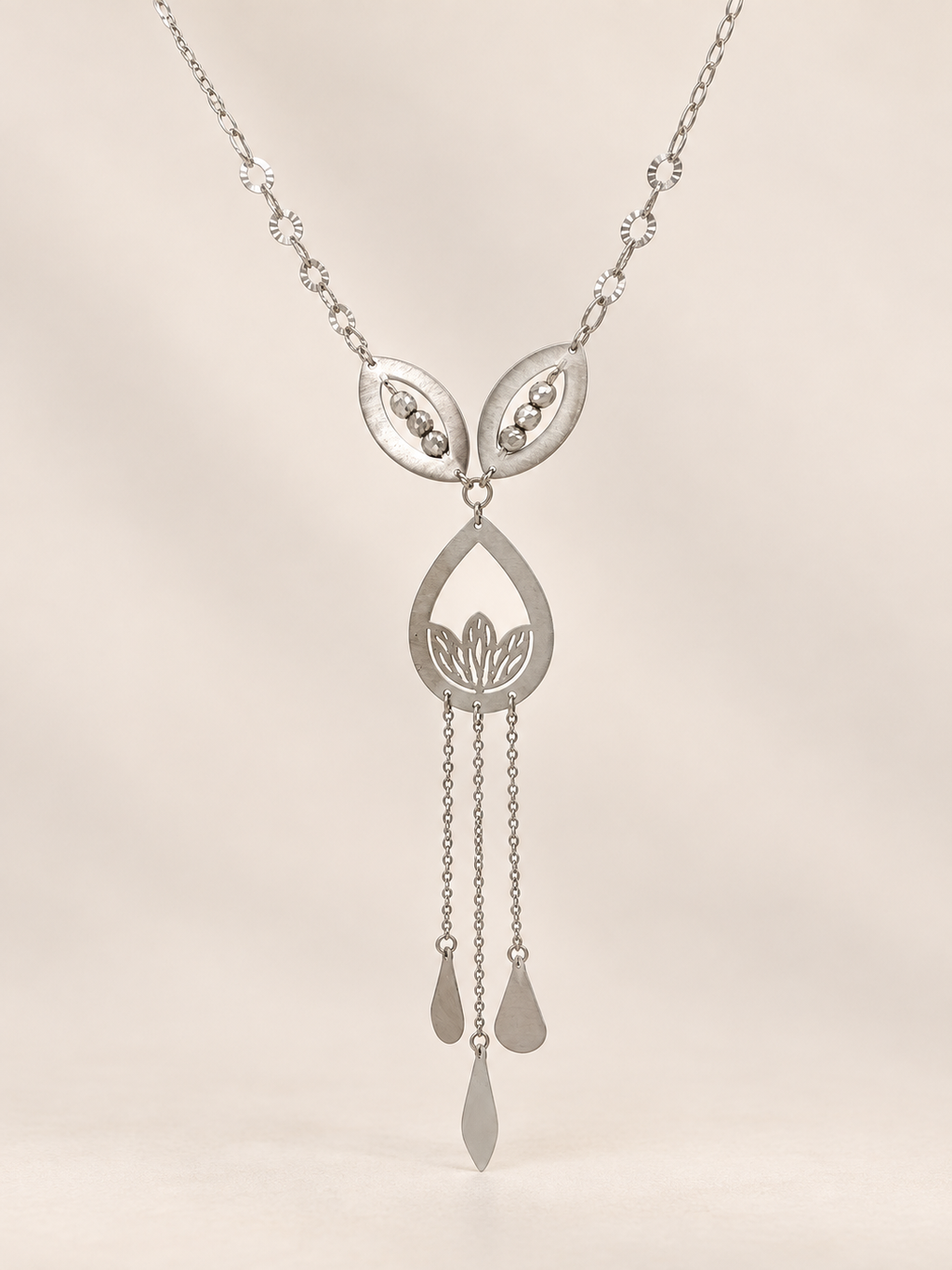 A silver-toned pendant necklace featuring a teardrop-shaped central pendant with a lotus motif, flanked by two leaf-like elements, with three dangling teardrop chains below, shown against a soft beige background.