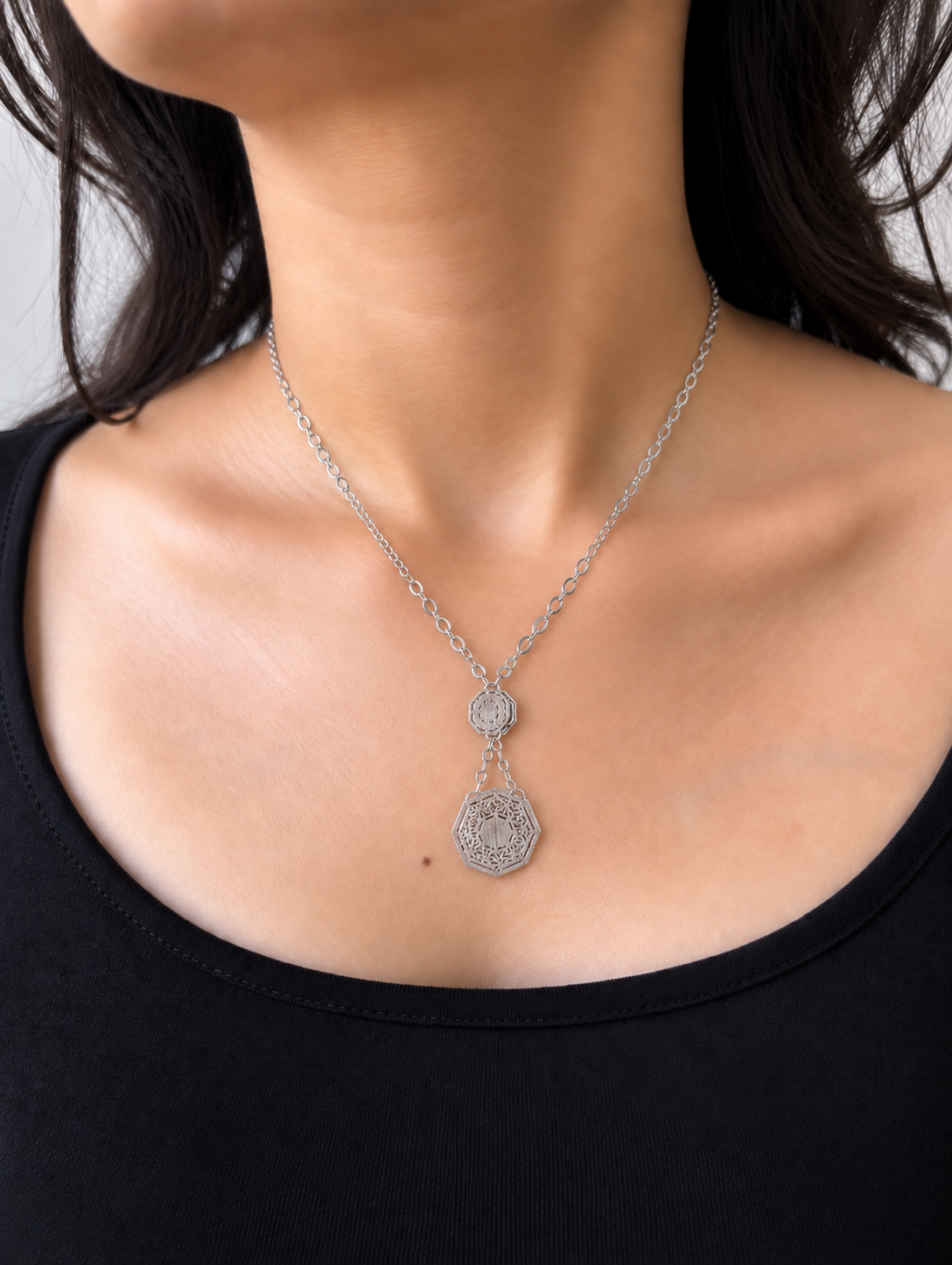 A silver-toned necklace featuring a delicate chain and a two-tiered pendant design, with a larger octagonal pendant hanging below a smaller circular one, worn by a model with a black top.