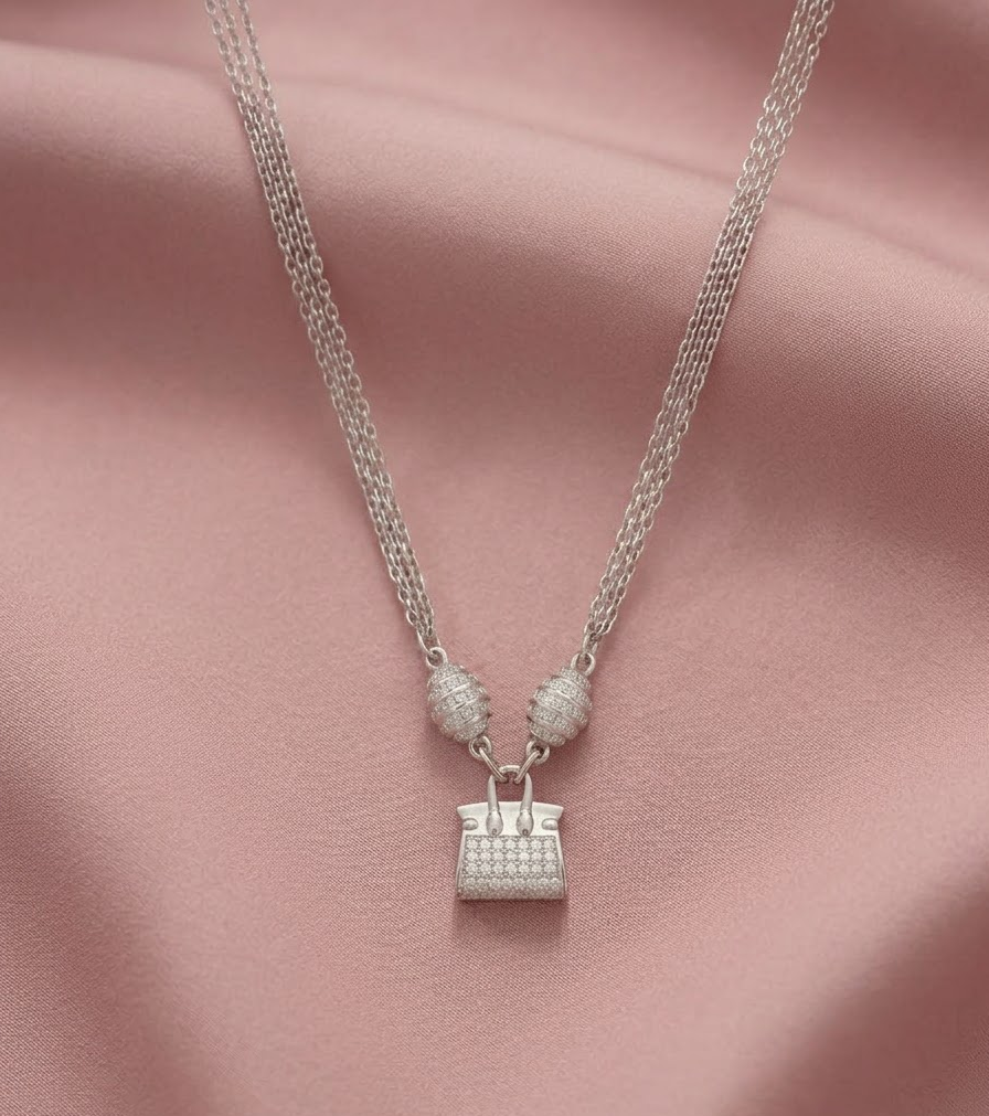 A silver-toned necklace featuring a small handbag-shaped pendant and two round textured beads, displayed on draped pink satin fabric.