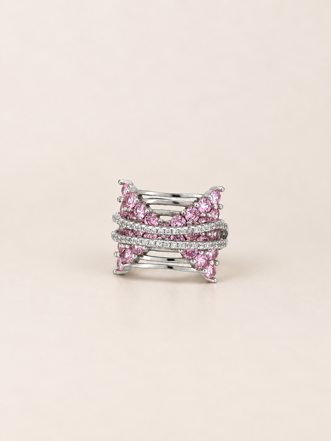 A silver-toned multi-band ring featuring rows of pink gemstones and pavé-set clear stones, photographed on a solid light beige background.