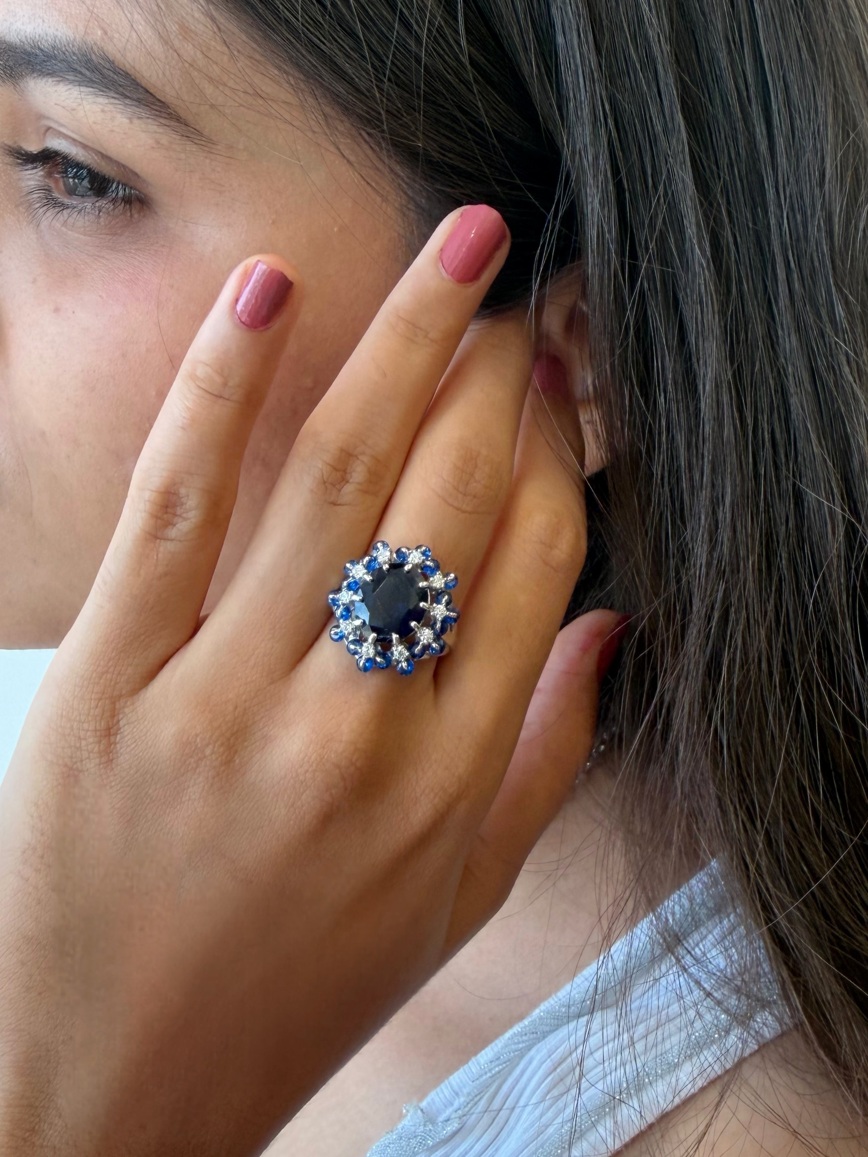 A close-up lifestyle shot of a woman wearing a ring with a dark blue oval center stone surrounded by a halo of blue and clear stones on her hand.