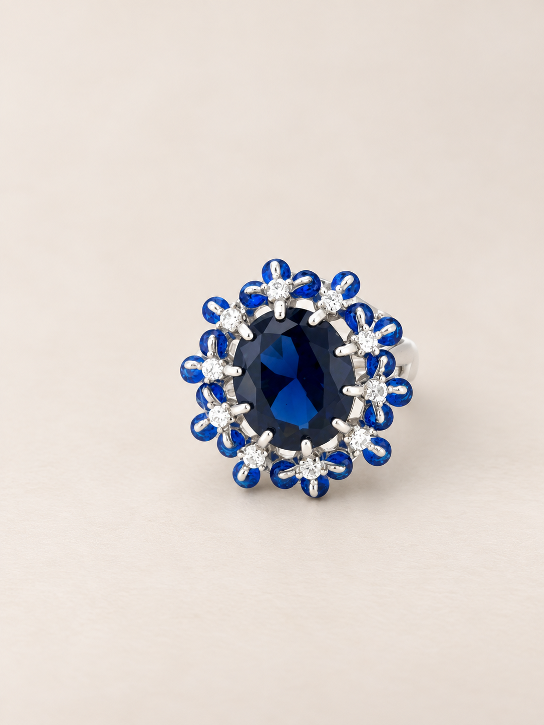 A silver ring featuring a large oval deep blue center stone surrounded by a halo of small blue stones and clear accent stones, set against a plain beige background.