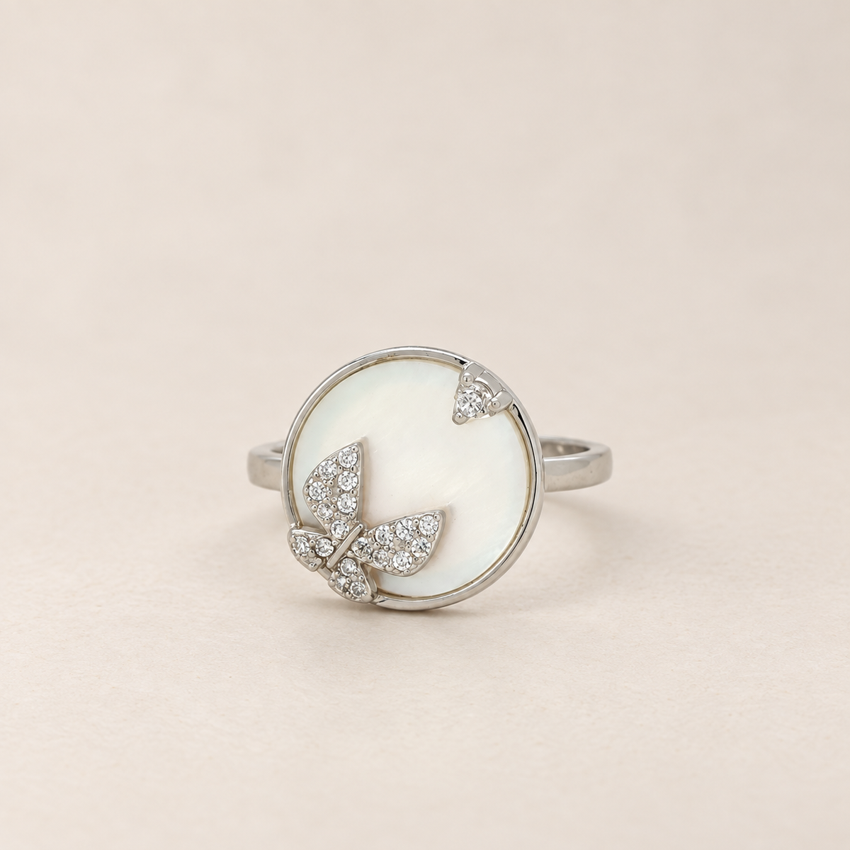 A silver ring featuring a round white mother-of-pearl inlay with a butterfly motif set with small clear crystals, photographed on a neutral beige background.