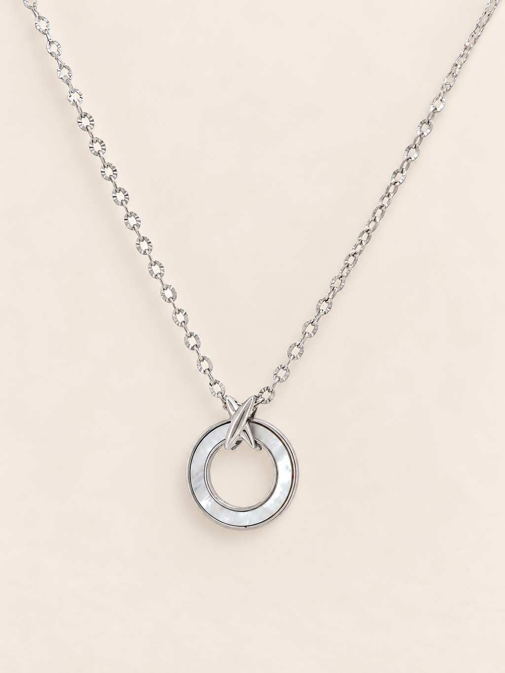 A silver-toned necklace featuring a delicate chain and a circle pendant with a polished finish, displayed against a solid light beige background.