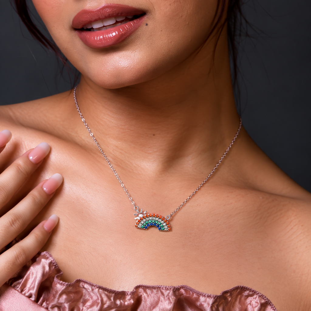A silver chain necklace featuring a small rainbow charm paved with multicolored rhinestones and a tiny silver star accent, worn by a model.