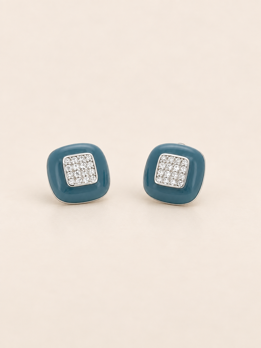 A pair of square stud earrings with a teal blue enamel-like border and a silver-toned square center paved with clear stones, set against a solid light beige background.