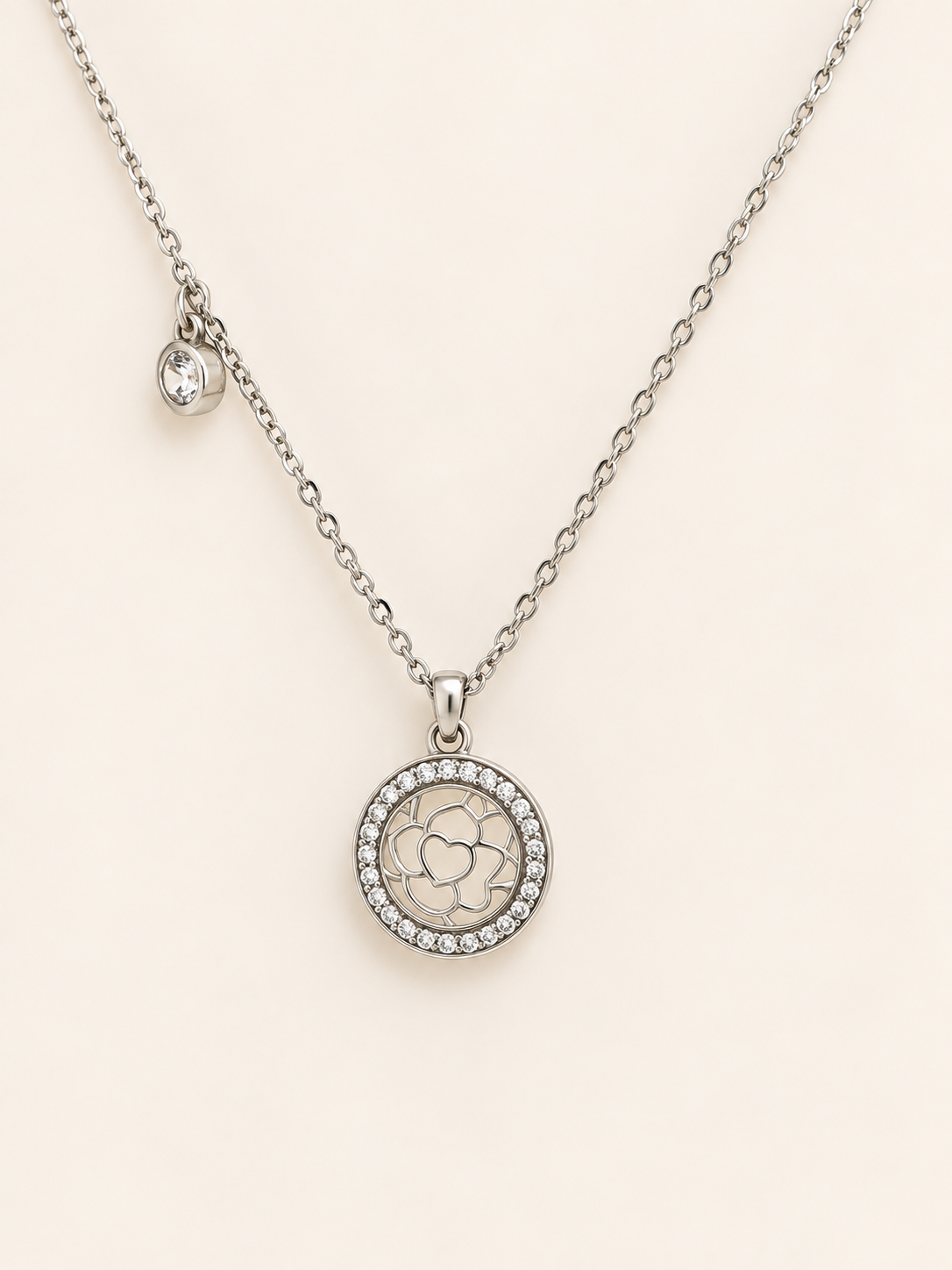 A silver-toned necklace featuring a circular pendant with a floral engraving and a border of small clear stones, displayed on a soft cream background.