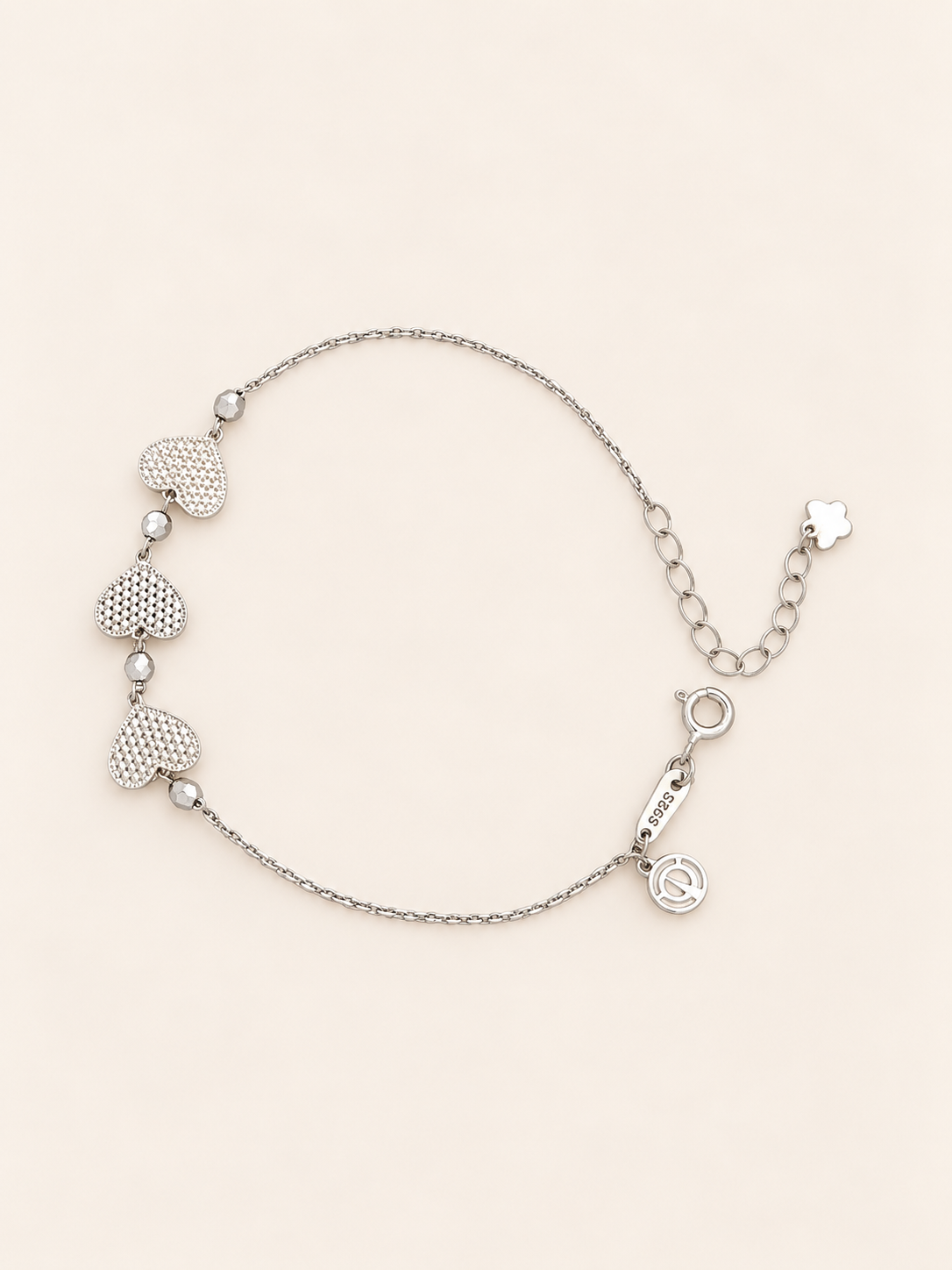A delicate silver chain bracelet featuring three textured heart charms, small round beads, displayed on a light beige background.