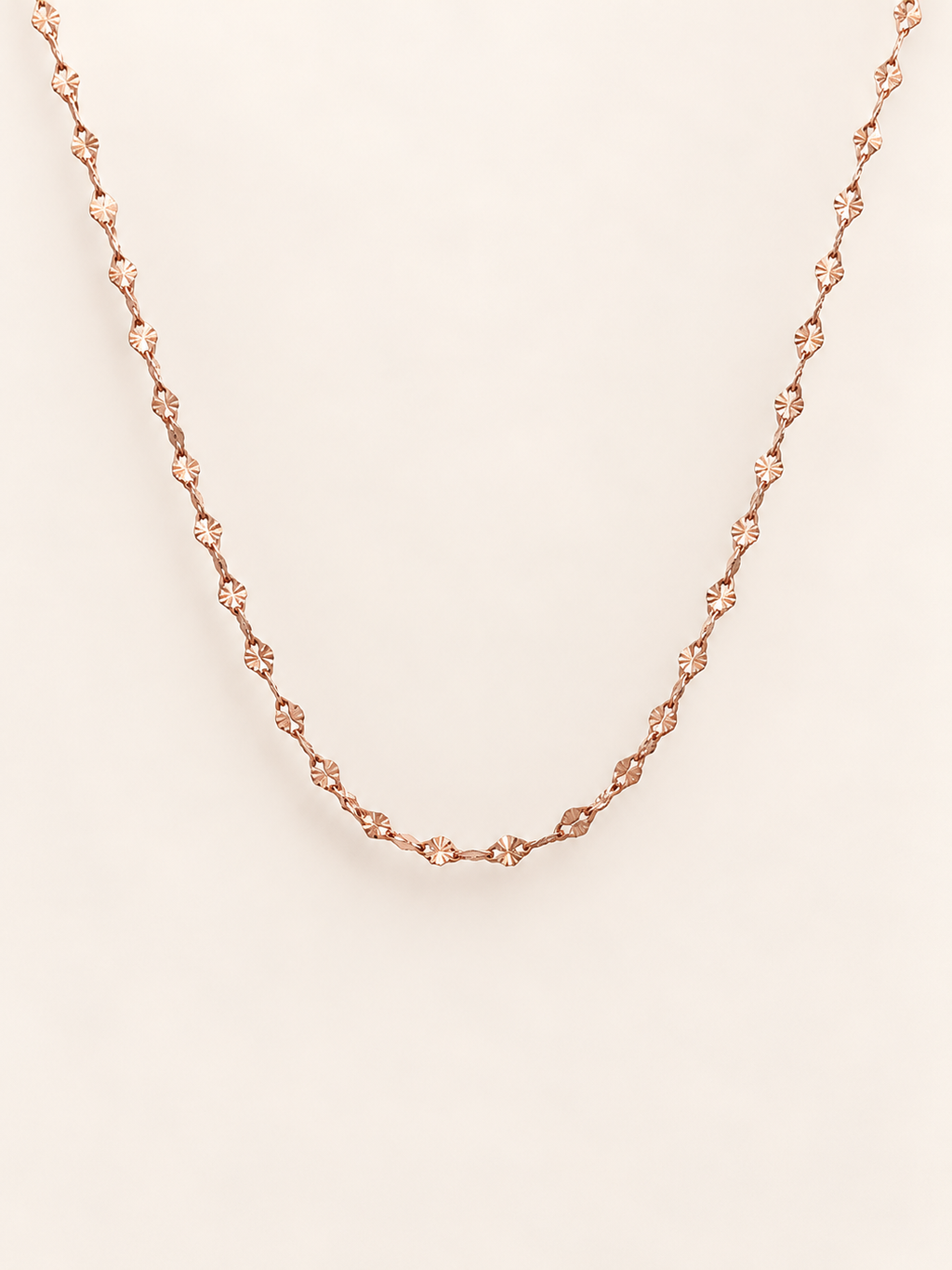 A delicate rose gold chain necklace, displayed against a solid, warm off-white background.