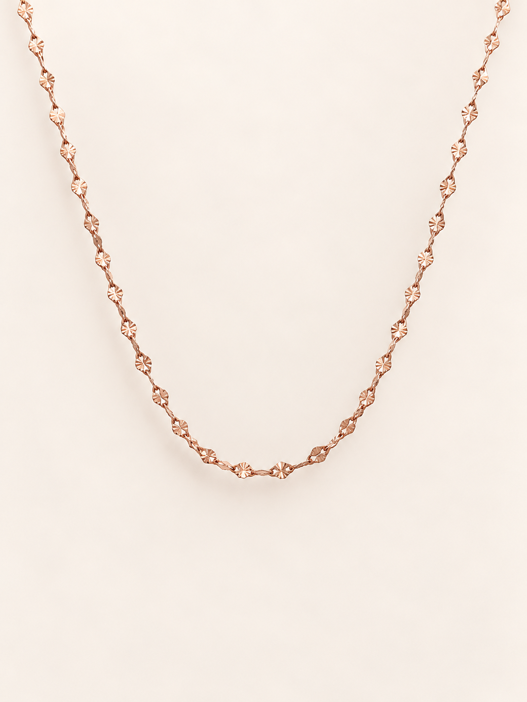 A delicate rose gold chain necklace, displayed against a solid, warm off-white background.