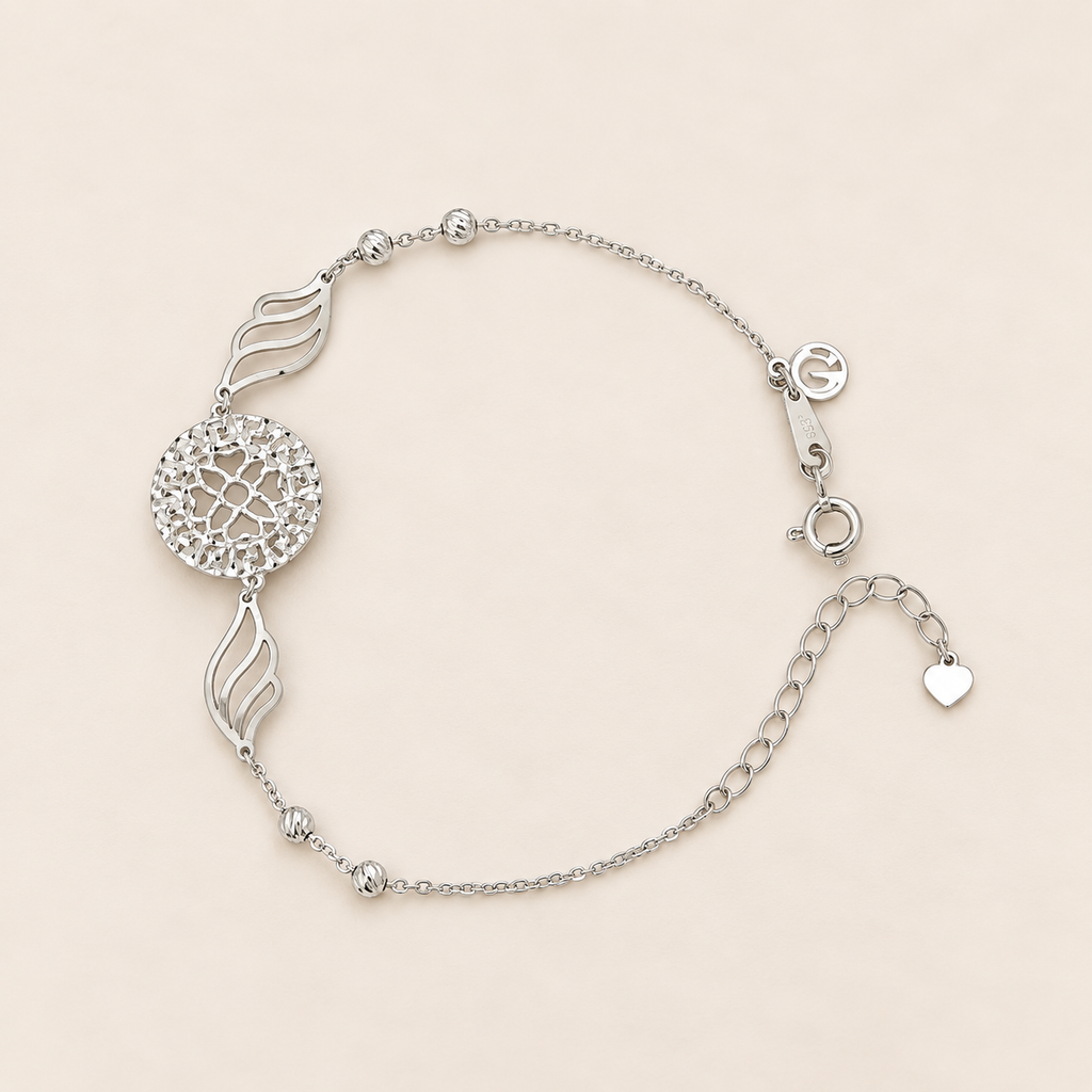 A silver dreamcatcher charm bracelet with a delicate chain, featuring a central circular woven web design, two flame-shaped accents, and a small heart charm, displayed on a light beige background.