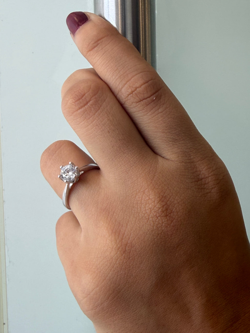A close-up of a hand wearing a silver solitaire engagement ring with a round clear gemstone, resting against a pale background.