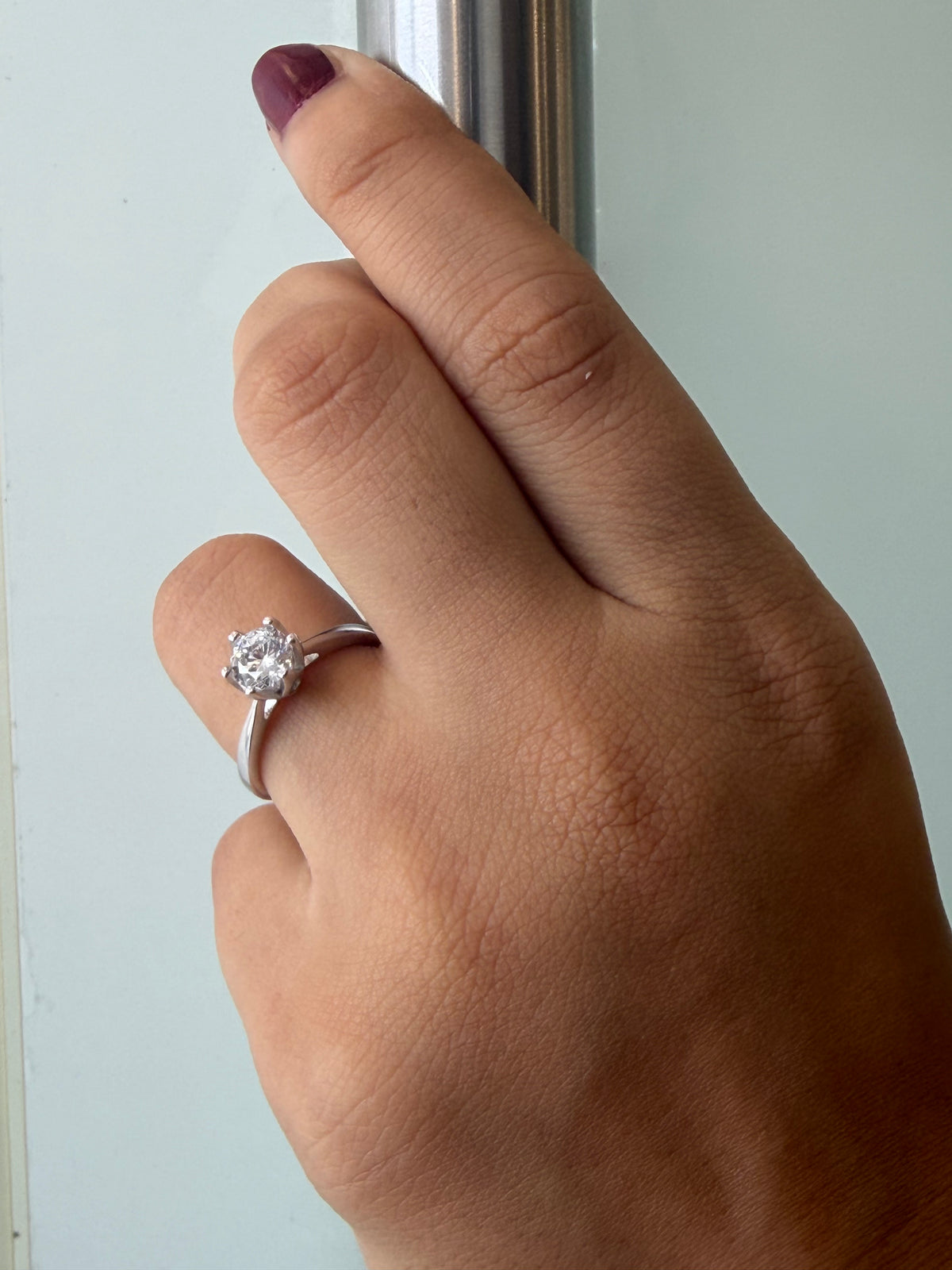 A close-up of a hand wearing a silver solitaire engagement ring with a round clear gemstone, resting against a pale background.