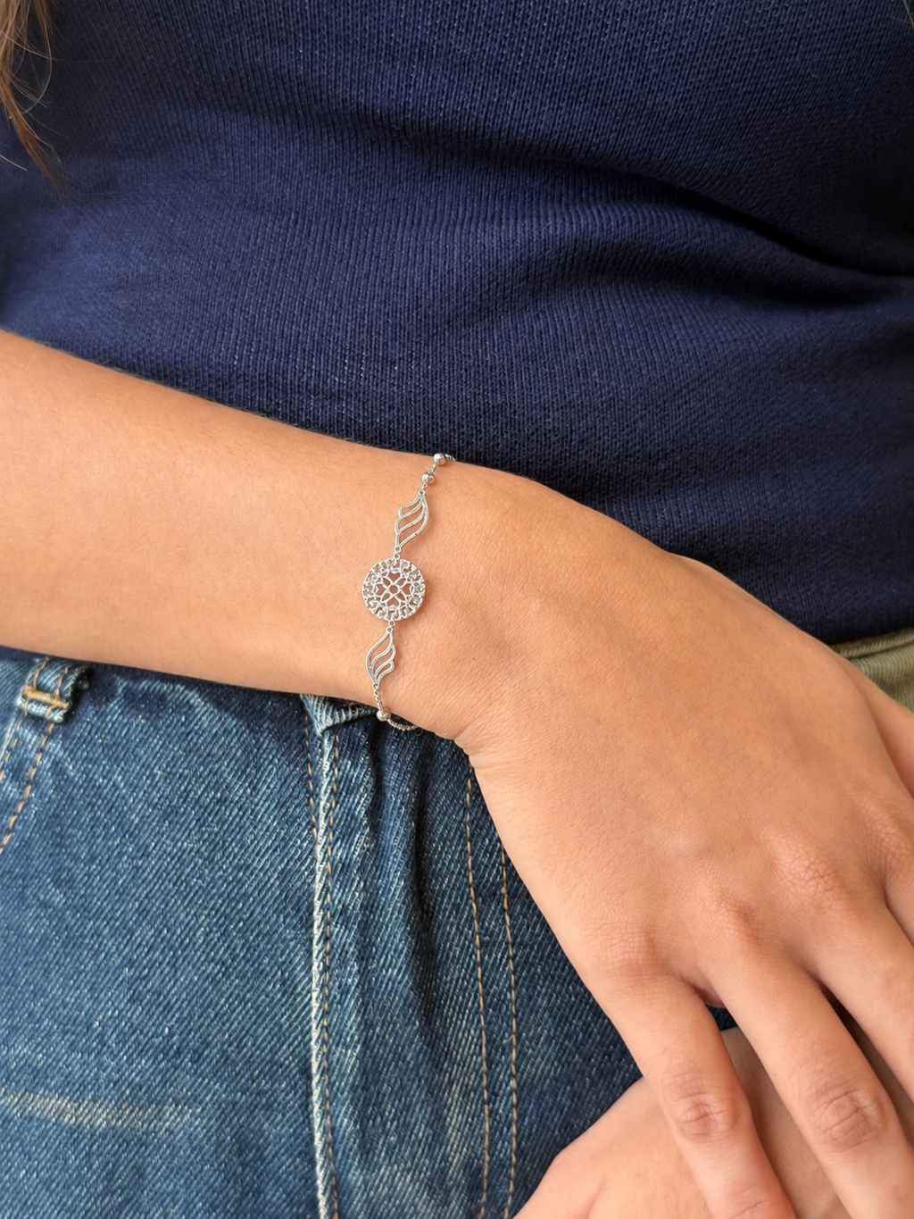 A close-up of a person's wrist wearing a delicate silver bracelet with a circular sunburst center and two wavy side links, styled with a navy blue knit top and blue jeans.