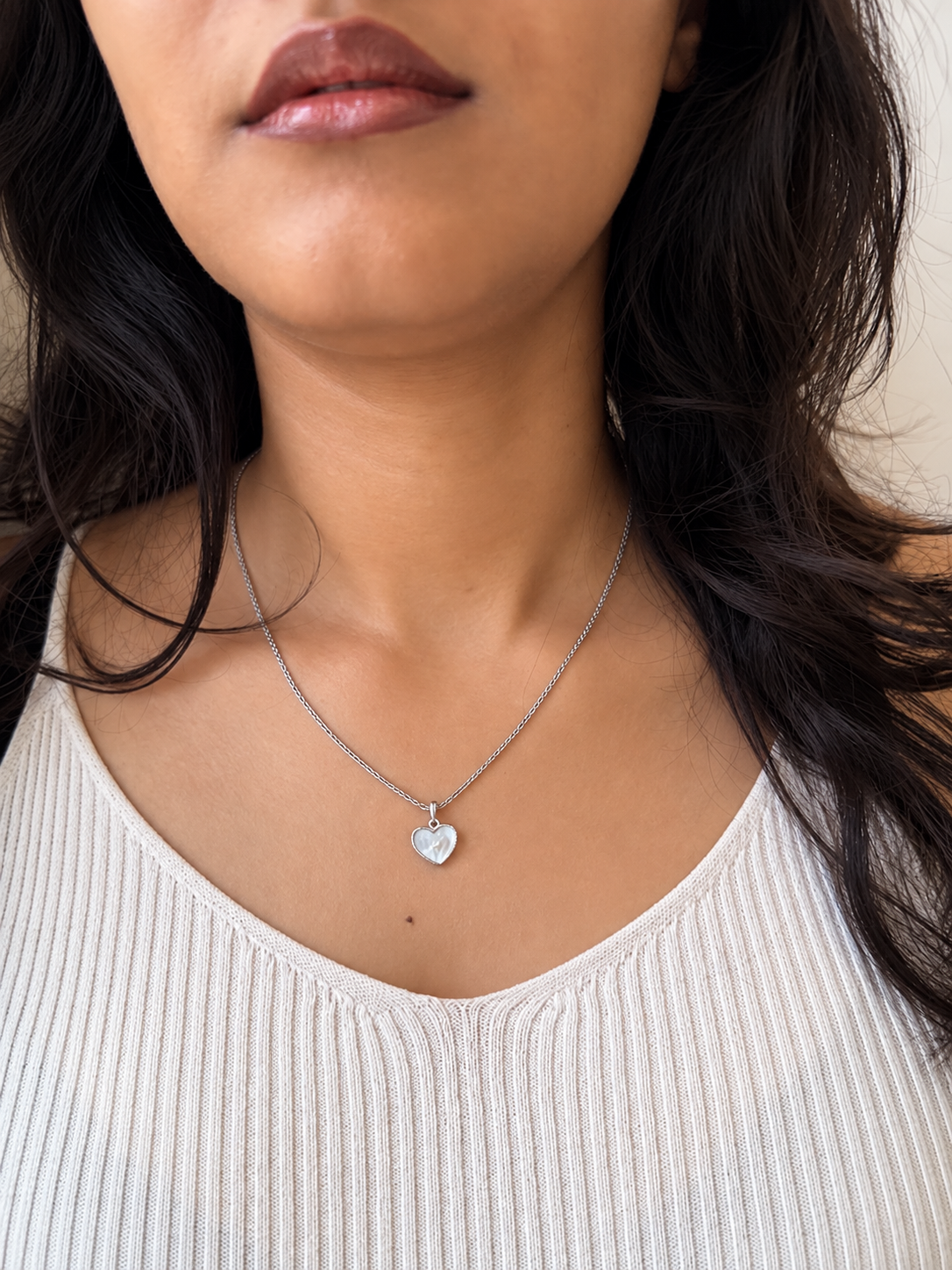A close-up of a woman wearing a delicate silver heart pendant necklace on a fine chain, paired with a ribbed cream-colored top.