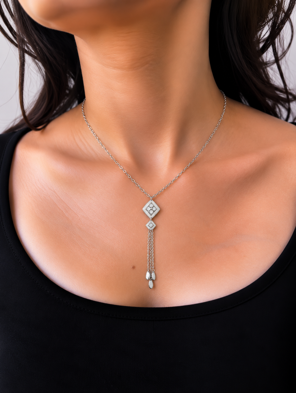 A silver-toned dangle necklace featuring a diamond-shaped pendant with a clear gemstone, a smaller diamond accent, and a dangling chain ending in a teardrop-shaped clear stone, worn by a model.