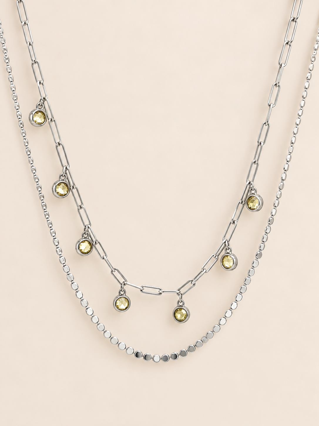 A layered silver necklace set featuring a paperclip chain, a beaded chain, and a delicate chain with small round yellow-green gemstone charms, displayed on a solid pale pink background.