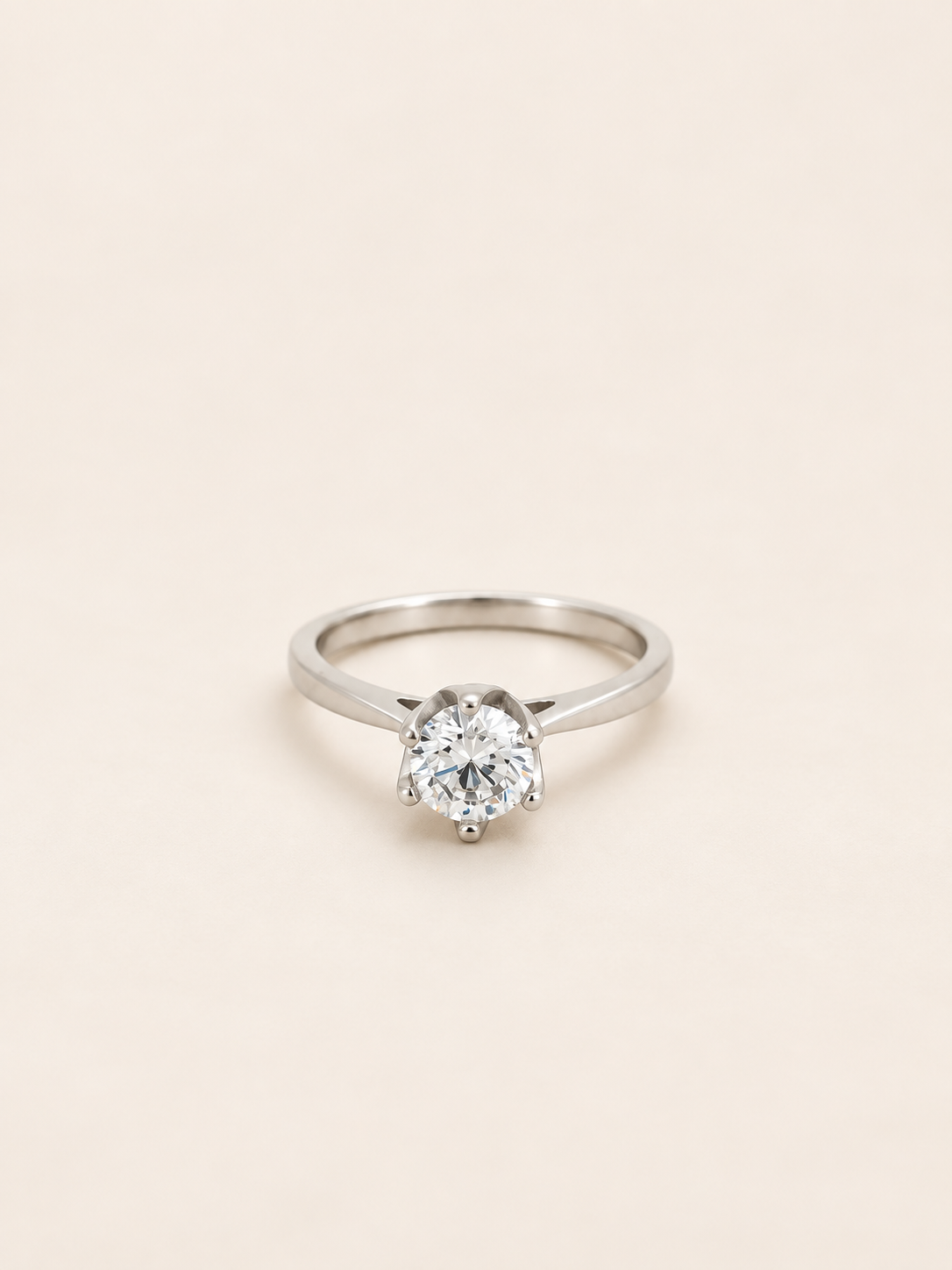 A classic solitaire engagement ring featuring a round brilliant-cut diamond set in a polished silver-colored band, presented on a solid, warm off-white background.