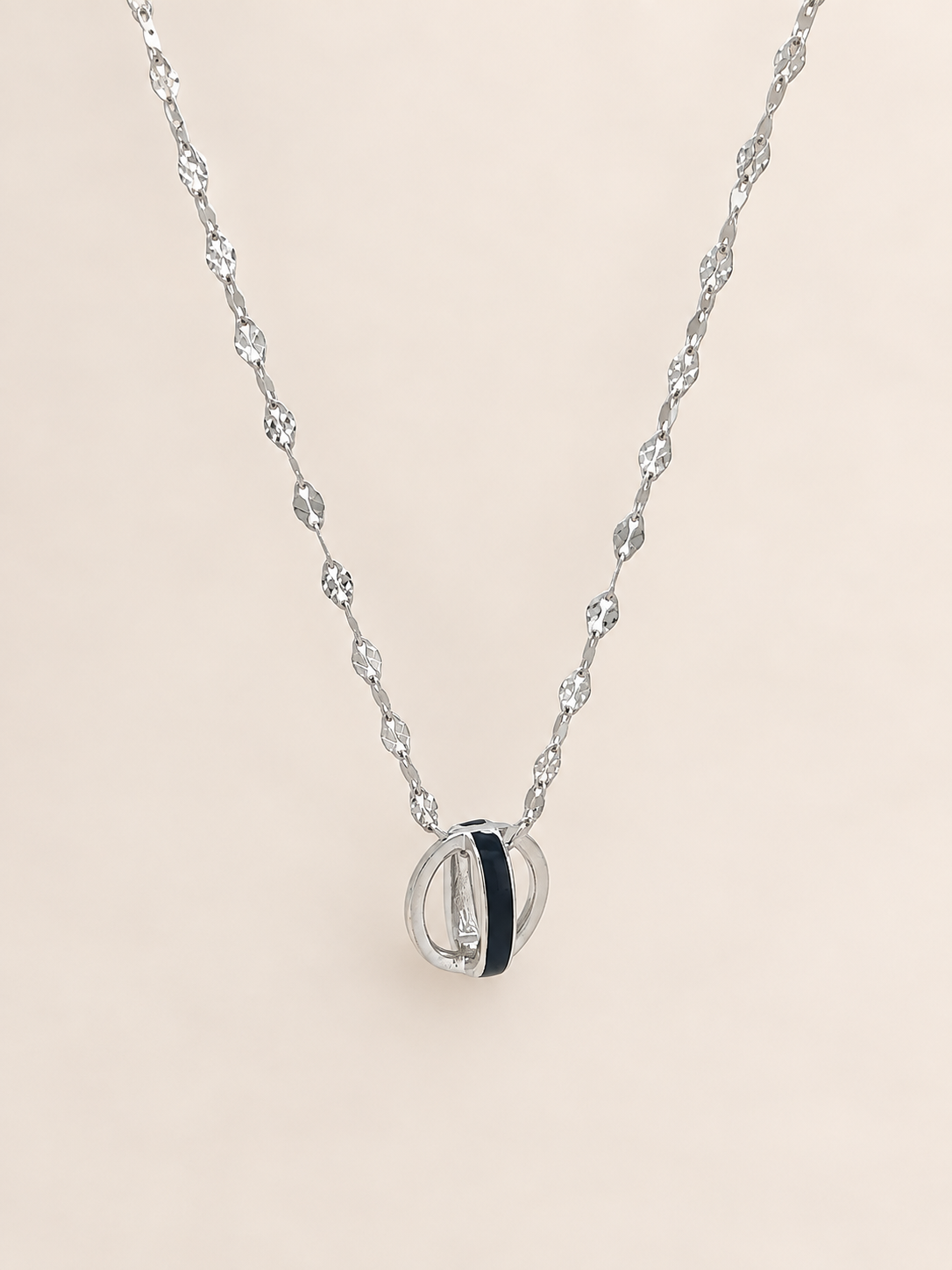 A silver-toned necklace with a delicate chain and a pendant featuring interlocking silver rings and a central black inlay, displayed on a soft beige background.