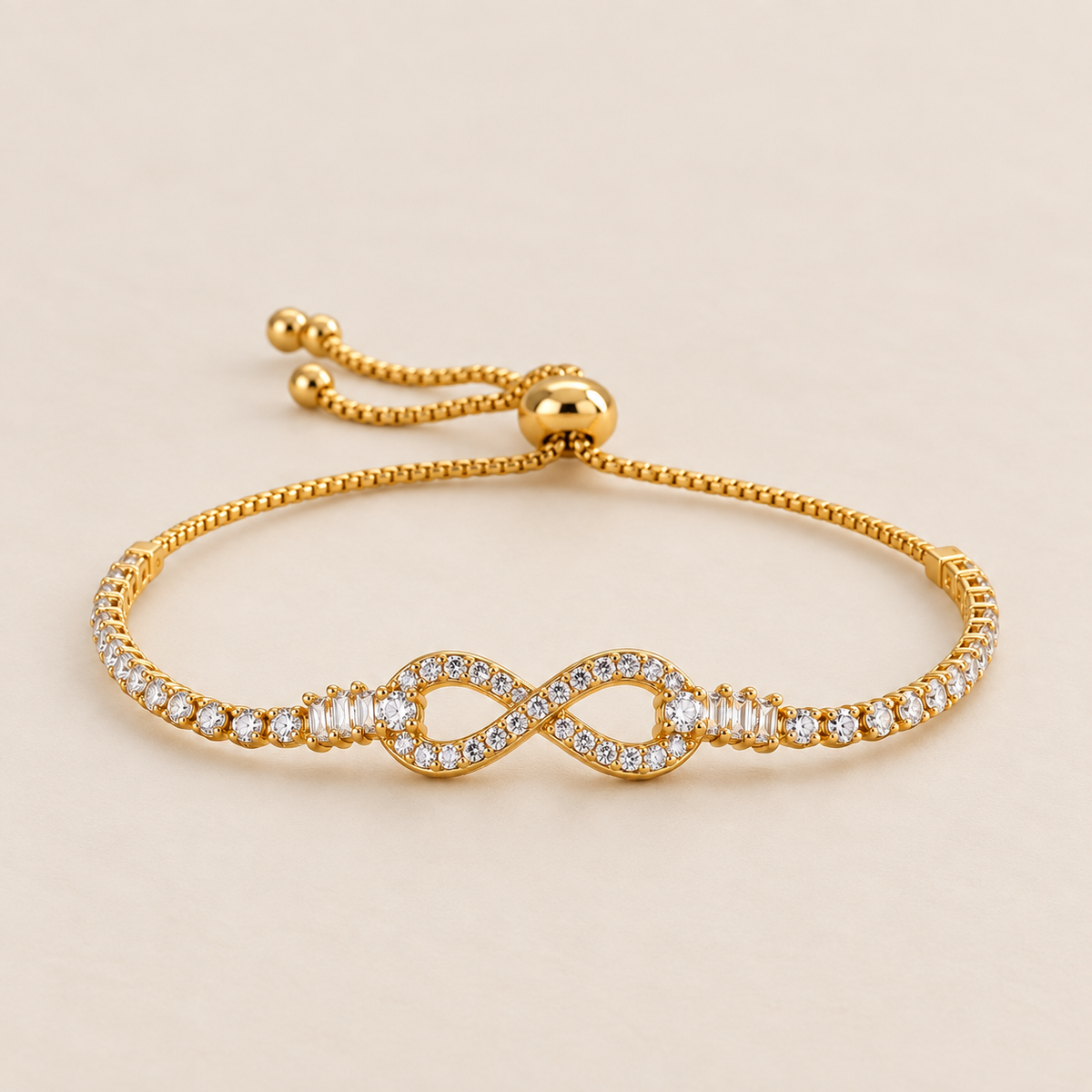 A delicate gold chain bracelet with a infinity design in the middle filled with diamonds and both sides has circle diamonds displayed against a light pink background.