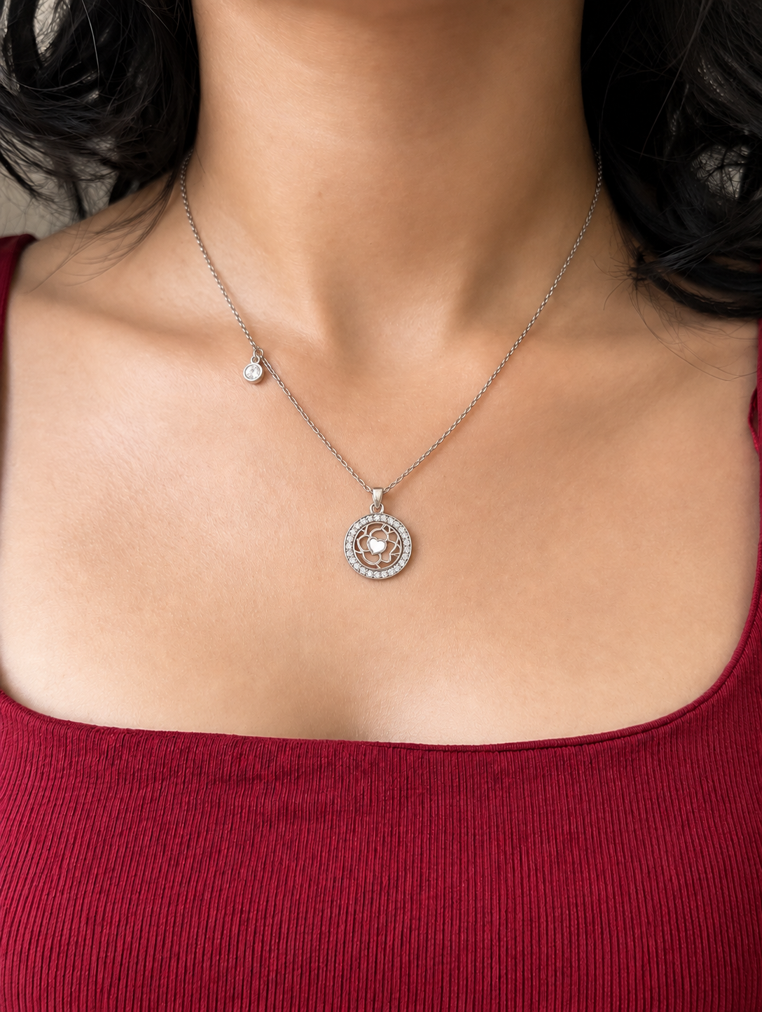 A close-up of a woman wearing a delicate silver chain necklace with a circular pendant featuring a central clear stone surrounded by small sparkling stones, which have a flower inside and a small dangle on the chain.