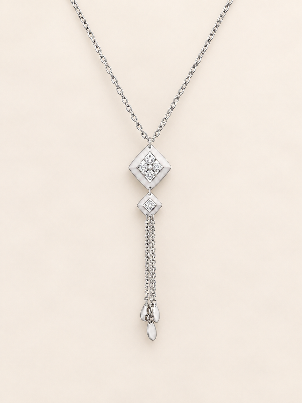A silver-toned dangle necklace featuring a diamond-shaped pendant with a clear gemstone, a smaller diamond accent, and a dangling chain ending in a teardrop-shaped clear stone, set against a solid pale beige background.