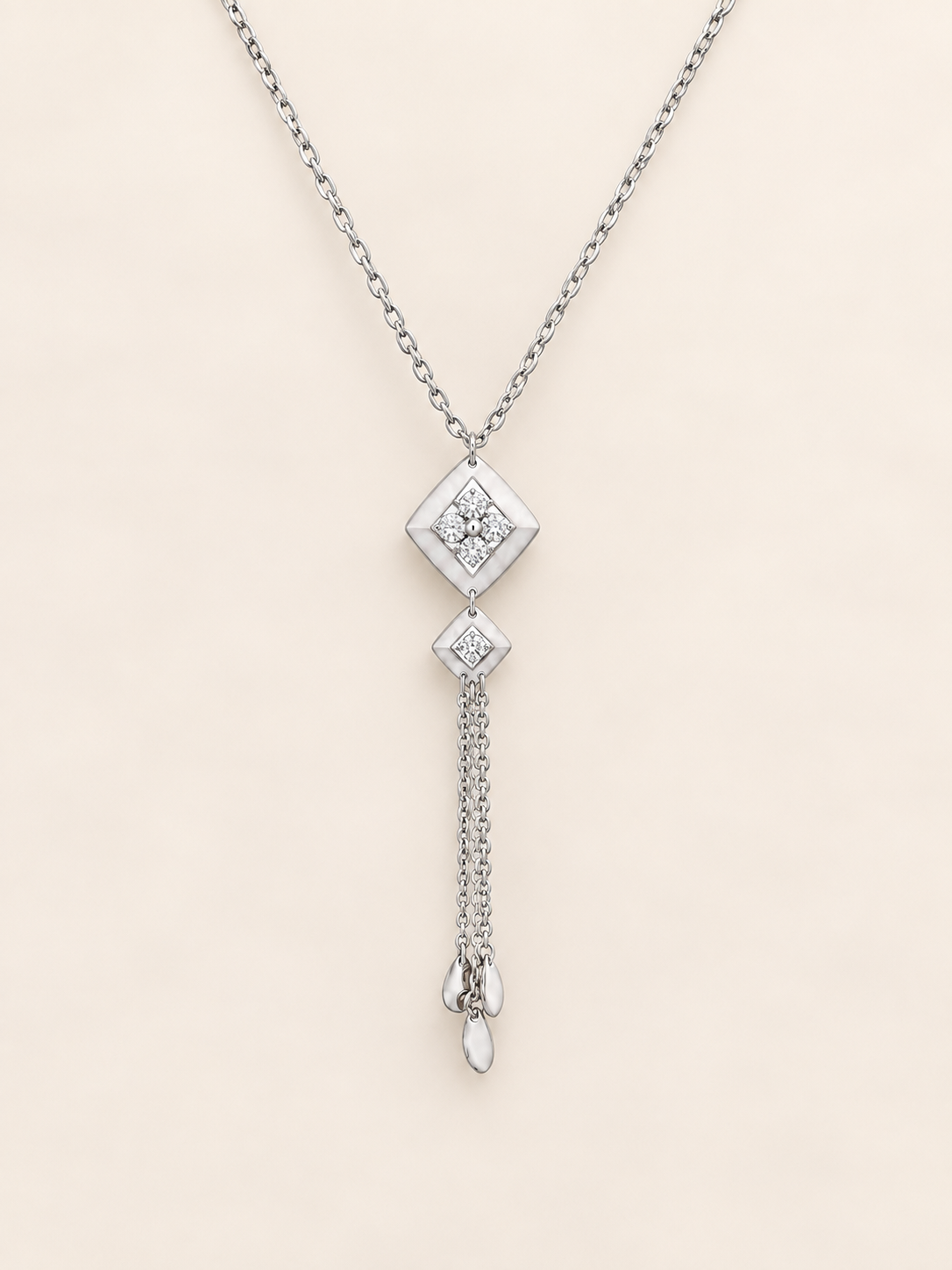 A silver-toned dangle necklace featuring a diamond-shaped pendant with a clear gemstone, a smaller diamond accent, and a dangling chain ending in a teardrop-shaped clear stone, set against a solid pale beige background.