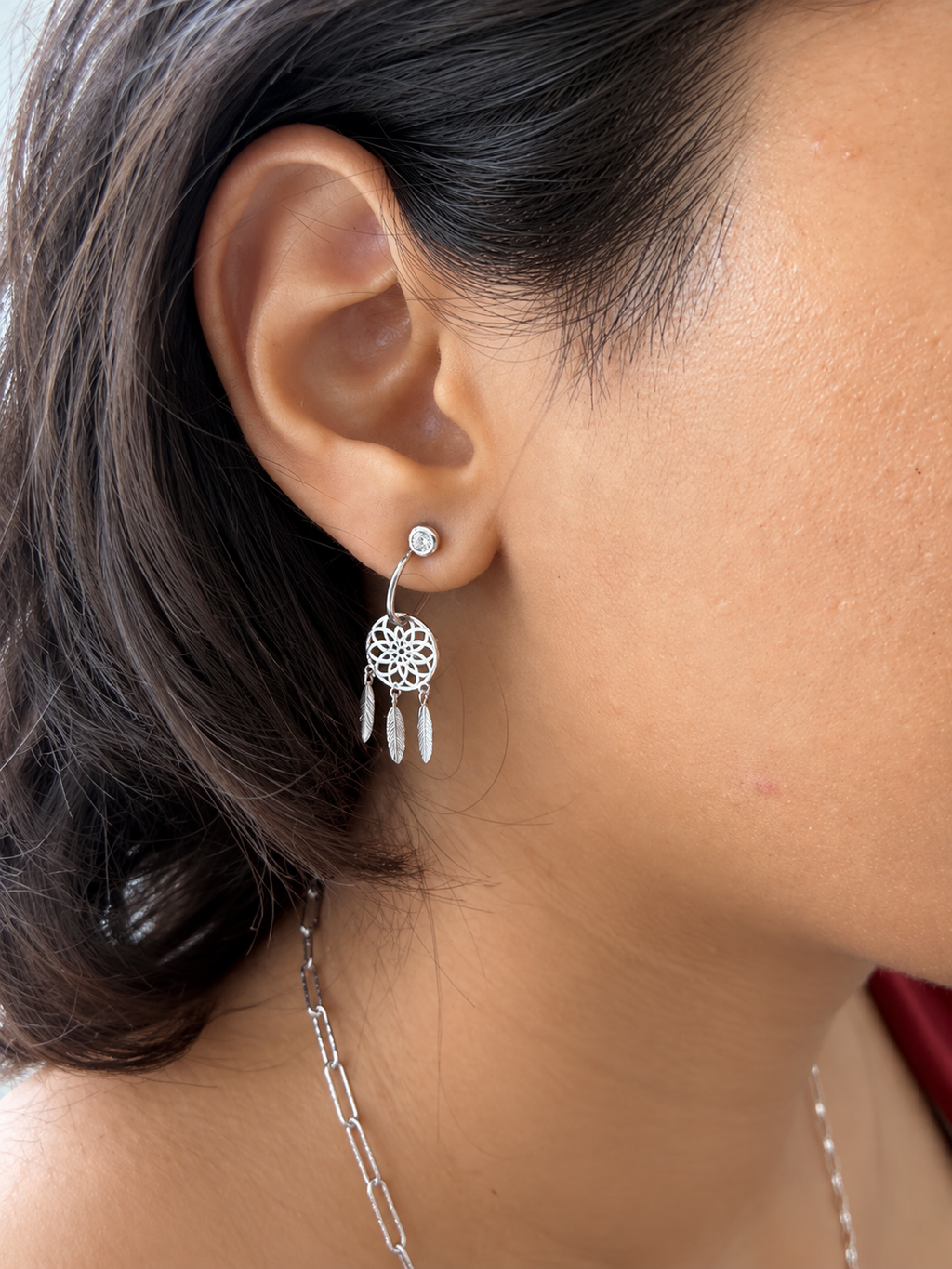 A close-up of a person's ear wearing a silver dreamcatcher dangle earring with three dangling feathers and a small clear stone stud.