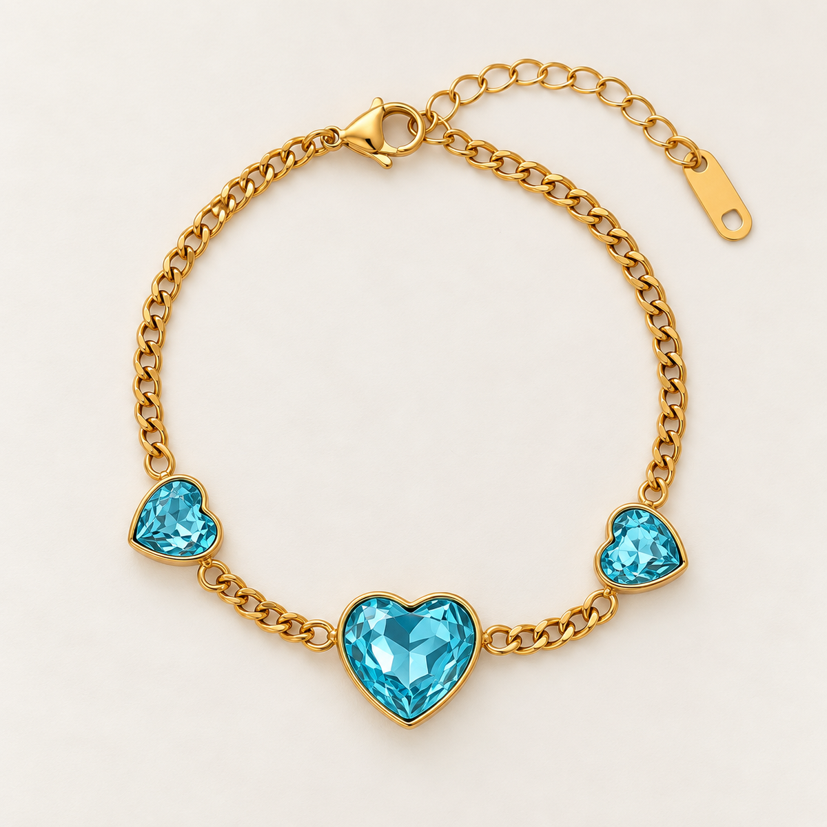 Gold chain bracelet with three heart-shaped turquoise blue crystals, displayed on a clean light pink background.