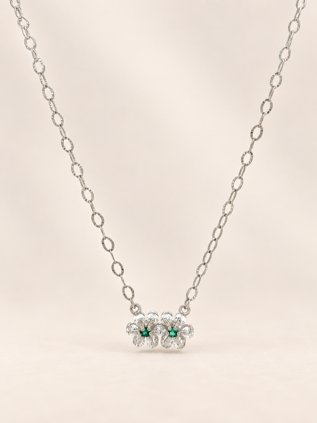 A silver chain necklace featuring a pendant of three small flower clusters, each with a central green gemstone surrounded by clear stones, set against a soft beige background.