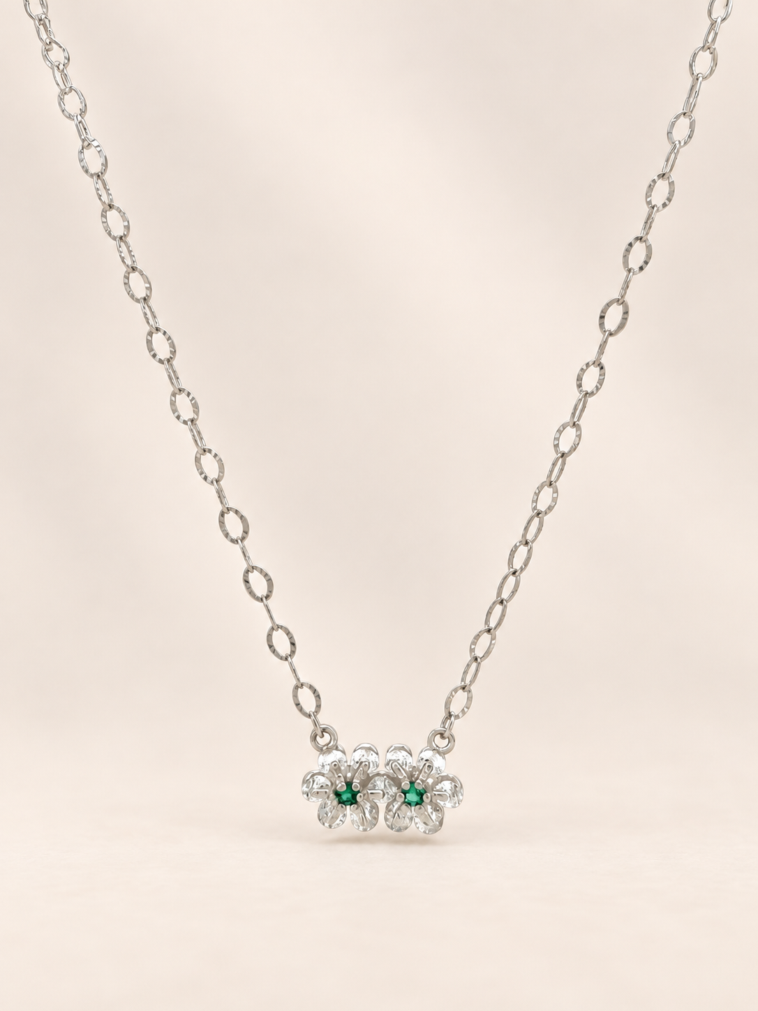 A silver chain necklace featuring a pendant of three small flower clusters, each with a central green gemstone surrounded by clear stones, set against a soft beige background.