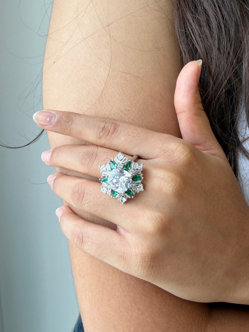 A close-up of a hand wearing a large floral cluster ring with a central clear gemstone and surrounding green and silver stones, shown on a person's wrist against a soft gray background.