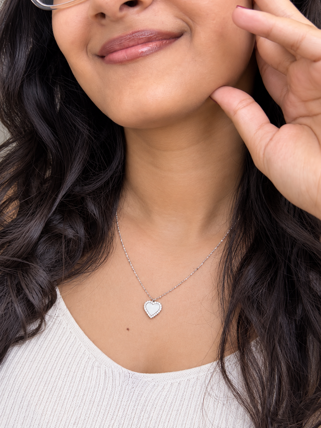 A close-up lifestyle shot of a woman wearing a delicate silver heart pendant necklace on a fine chain, paired with a white ribbed top.