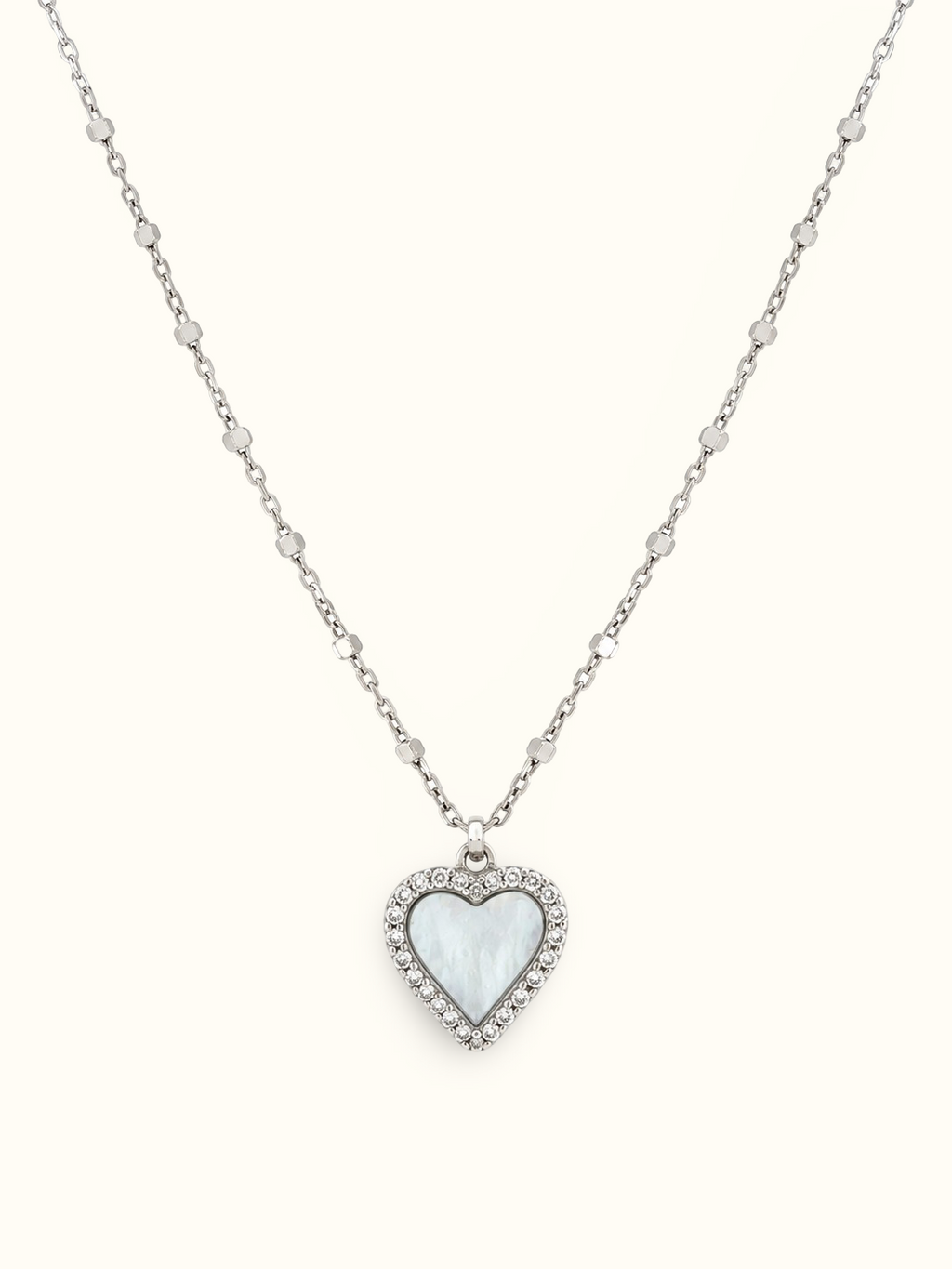 A silver heart pendant necklace featuring a milky white mother-of-pearl heart inlay, surrounded by a halo of small clear crystals on a delicate silver chain, set against a solid off-white background.